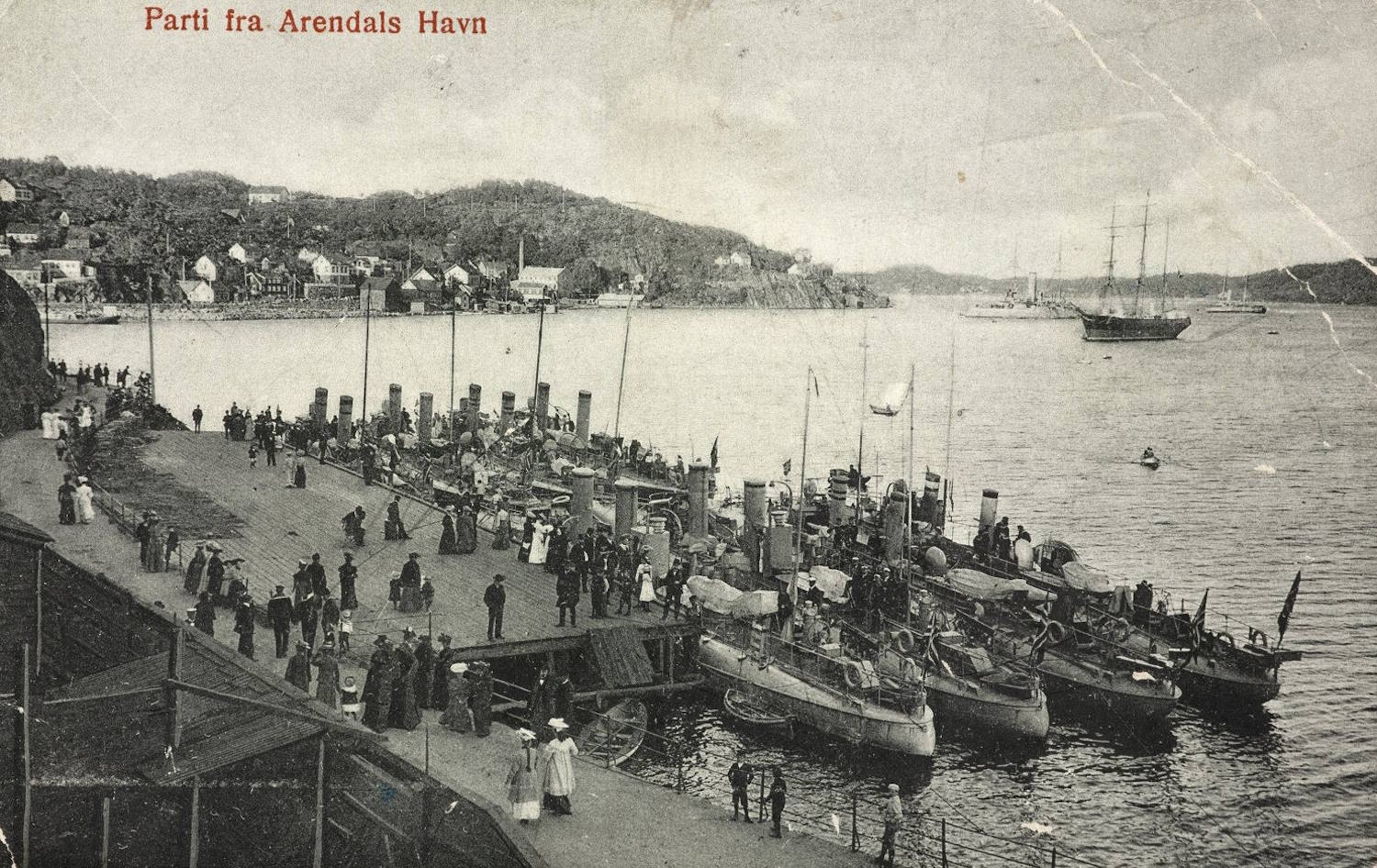 Flåtebesøk i Arendal 1905. Bildet viser en travel brygge i en havn, sett litt ovenfra og på skrå. Mange mennesker står og går rundt på den brede kaien. De fleste er kledd i tidstypiske klær fra tidlig 1900‑tall, inkludert lange kjoler, hatter og mørke frakker. Noen få barn kan også sees. Langs bryggen ligger en rekke mindre båter tett i tett, de fleste med master og rigg. Båtene virker å være små fartøy eller patruljebåter av eldre type, og flere har dekk fulle av utstyr. På sjøen utenfor ligger to større seilskip ankret opp, med høye master og flere seil som er surret. I bakgrunnen vises en lav, kupert kystlinje med spredte hvite hus og skogkledde åser. Vannet i havnen ligger rolig. Øverst i bildet står teksten «Parti fra Arendals Havn», som indikerer at motivet er fra Arendal. Bildet er i sort-hvitt eller sepia og har preg av å være et gammelt postkort eller historisk fotografi.