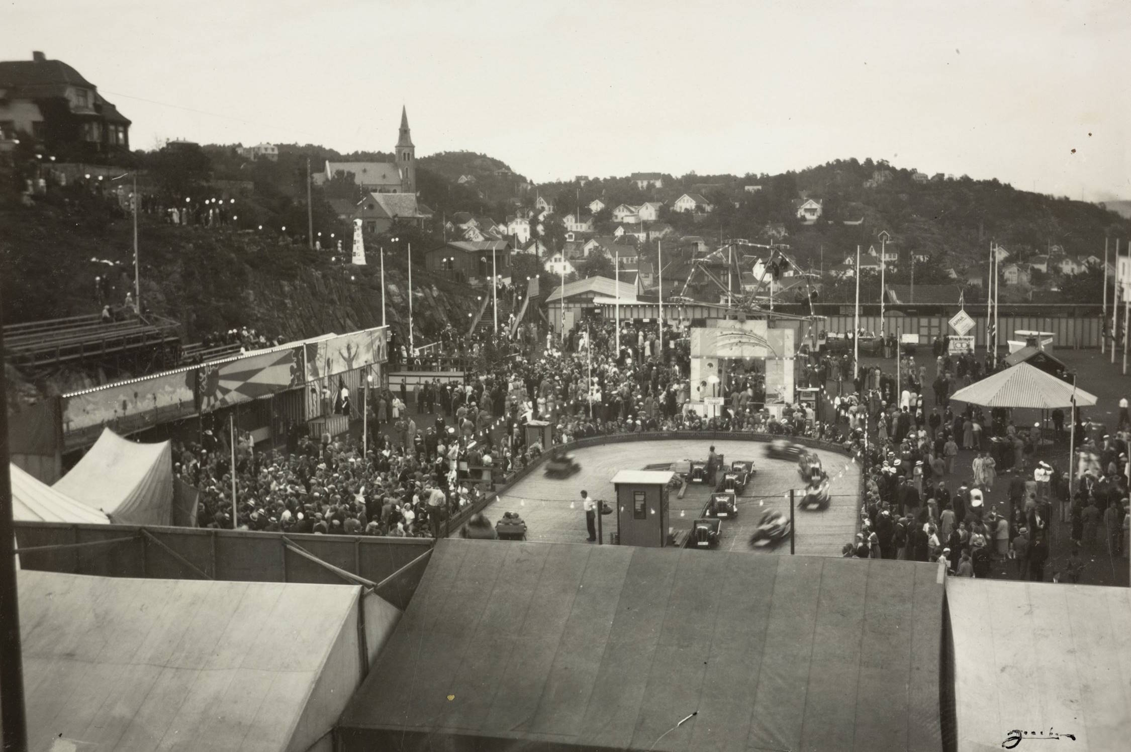Folksomt i fornøyelsesavdelingen på Idrettsplassen under varemessen i Arendal i 1936. Foto: Jacobsen. Bildet viser et stort folkeliv på et utendørs messeområde. Foran i bildet ligger flere store telt med skrå tak. Bak teltene ser man et område med mange mennesker som beveger seg mellom boder, attraksjoner og stands. Midt i motivet ligger en rund kjørebane for små elektriske biler, der flere små kjøretøy er i bevegelse. Rundt banen står og går mange besøkende. Til høyre for banen skimtes et rundt bygg eller telt med stripet tak, sannsynligvis en annen attraksjon. I bakgrunnen strekker det seg en lang rekke boder og konstruksjoner bortover mot venstre. Mange flaggstenger står spredt gjennom området, noen med flagg heist. Folkemengden er tett og fyller store deler av plassen. Bak messeområdet ligger en bratt skråning med gjerde og sittebenker eller tribuner. Øverst på høyden og utover i horisonten sees en kirke med spiss tårn og mange trehus spredt i det kuperte landskapet. Hele scenen fremstår som en travel og livlig dag i fornøyelsesavdelingen på en varemessse.
