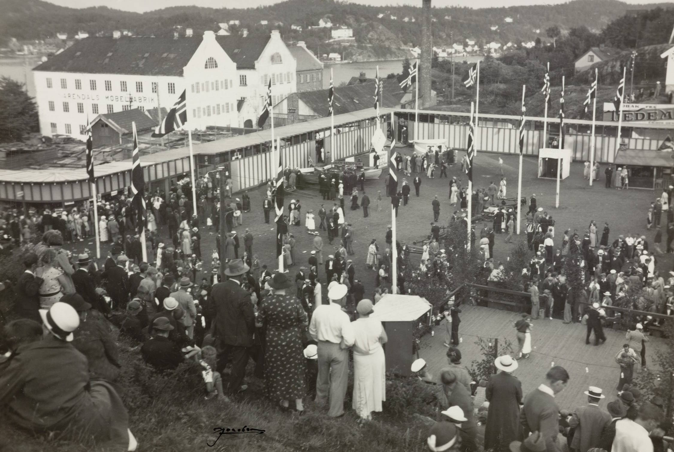 Idrettsplassen under varemessen i Arendal i 1936. Foto: Jacobsen. Bildet viser et stort uteområde fylt med mennesker som har samlet seg på en messe- eller festivalplass. Bildet er tatt fra en forhøyning, slik at man ser ned mot plassen. I forgrunnen står mange mennesker tett sammen på en gressbakke. De er vendt mot messeområdet og ser på aktivitetene under. De fleste er kledd i 1930‑tallsklær: lange frakker, kjoler, hatter og mørke dresser. Midt i bildet ligger et stort inngjerdet område med jevnt underlag. Her går og står mange mennesker i grupper, mens andre beveger seg mellom boder og små konstruksjoner. Rundt området står det en rekke flaggstenger med heiste norske flagg. Flere små telt eller boder står tett langs kanten av plassen. I bakgrunnen, på den andre siden av vannet eller en elv, ligger en hvit, stor bygning med markerte gavler. Bak denne sees mange mindre hus spredt i et kupert landskap med skogkledde åser. Stemningen er travel, som på en aktiv messedag med mange besøkende.