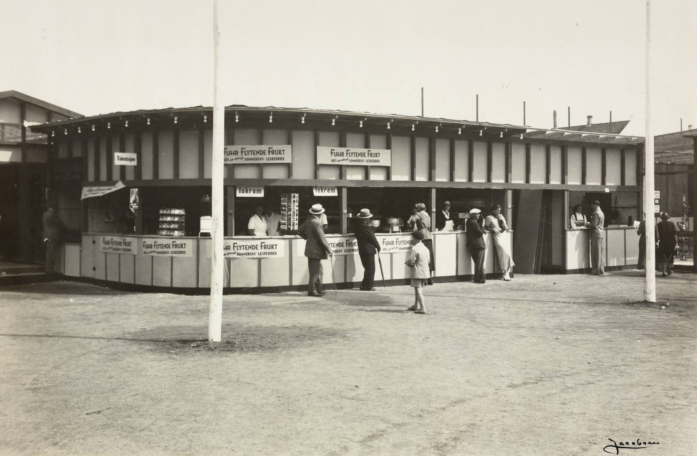 Kioskboder på Idrettsplassen under varemessen i Arendal i 1936. Foto: Jacobsen. Bildet viser et kiosk- og salgsområde på en messe eller markedsplass. Midt i motivet står en lang, lav bygning med flere åpne kioskutsalg vendt mot plassen. Bygningen har flatt tak med en rekke stolper som stikker opp langs kanten. Over flere av utsalgsdiskene henger skilt med teksten "Fuhr Flytende Frukt". I utsalgene ses flere personer som betjener kunder. Flere mennesker står foran bodene. Noen ser ut til å handle eller prate med ekspeditørene, mens andre bare står og betrakter området. De fleste er ikledd lyse eller mørke 1930‑tallsklær – menn med hatter og jakker, kvinner i kjoler og hodeplagg, og ett barn i lyst antrekk som står midt i plassen og ser mot bodene. I forgrunnen er det en åpen, flat plass, trolig grus- eller jorddekke. To flaggstenger står i bildet, én nær midten og én til høyre, begge uten flagg synlig i rammen. Bak kiosken skimtes andre små bygg og konstruksjoner, noe som antyder at dette er en del av et større messeområde.