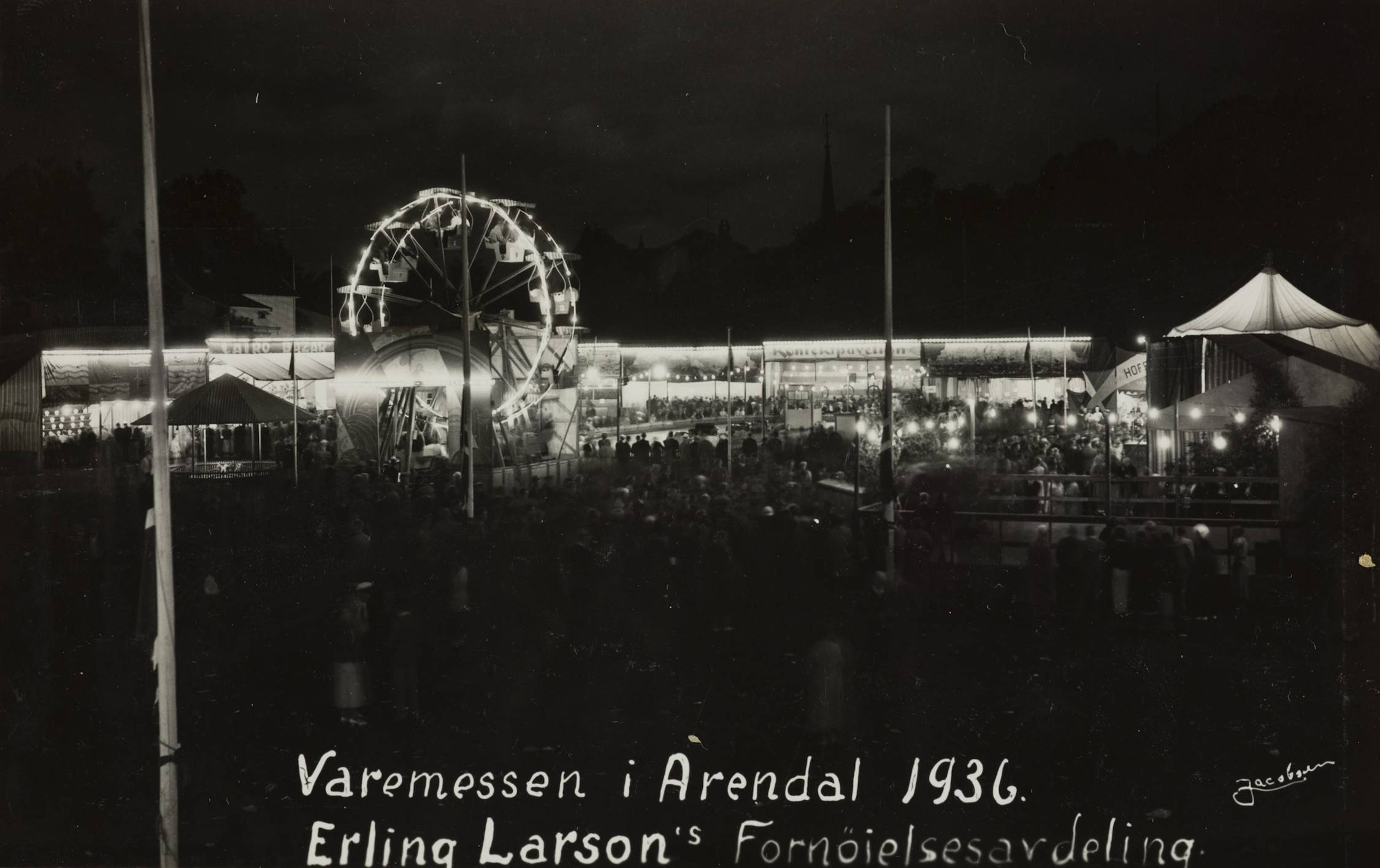Kveld i fornøyelsesavdelingen på Idrettsplassen under varemessen i Arendal i 1936. Foto: Jacobsen. Bildet viser et fornøyelsesområde på kveldstid under en messe. Det er mørkt, og lys fra attraksjoner og boder lyser opp scenen. Midt i bildet står et stort pariserhjul opplyst av mange små lyspærer som danner en klar sirkel mot den mørke himmelen. Rundt pariserhjulet ligger flere boder og små bygninger med sterke utendørslamper som lyser opp området. Det er mange mennesker på plassen, men de fleste fremstår som mørke silhuetter mot det sterke lyset fra attraksjonene. Til høyre står et telt med lyst, konisk tak, også opplyst. Langs kantene av plassen står flere flaggstenger. Noen flagg kan skimtes, men detaljene er vanskelig å se på grunn av mørket. Nederst i bildet, skrevet med hvit skrift, står teksten: "Varemessen i Arendal 1936 – Erling Larson's Fornøyelsesavdeling." Motivets helhet viser et livlig kveldsarrangement med lys, aktivitet og et stort antall besøkende.