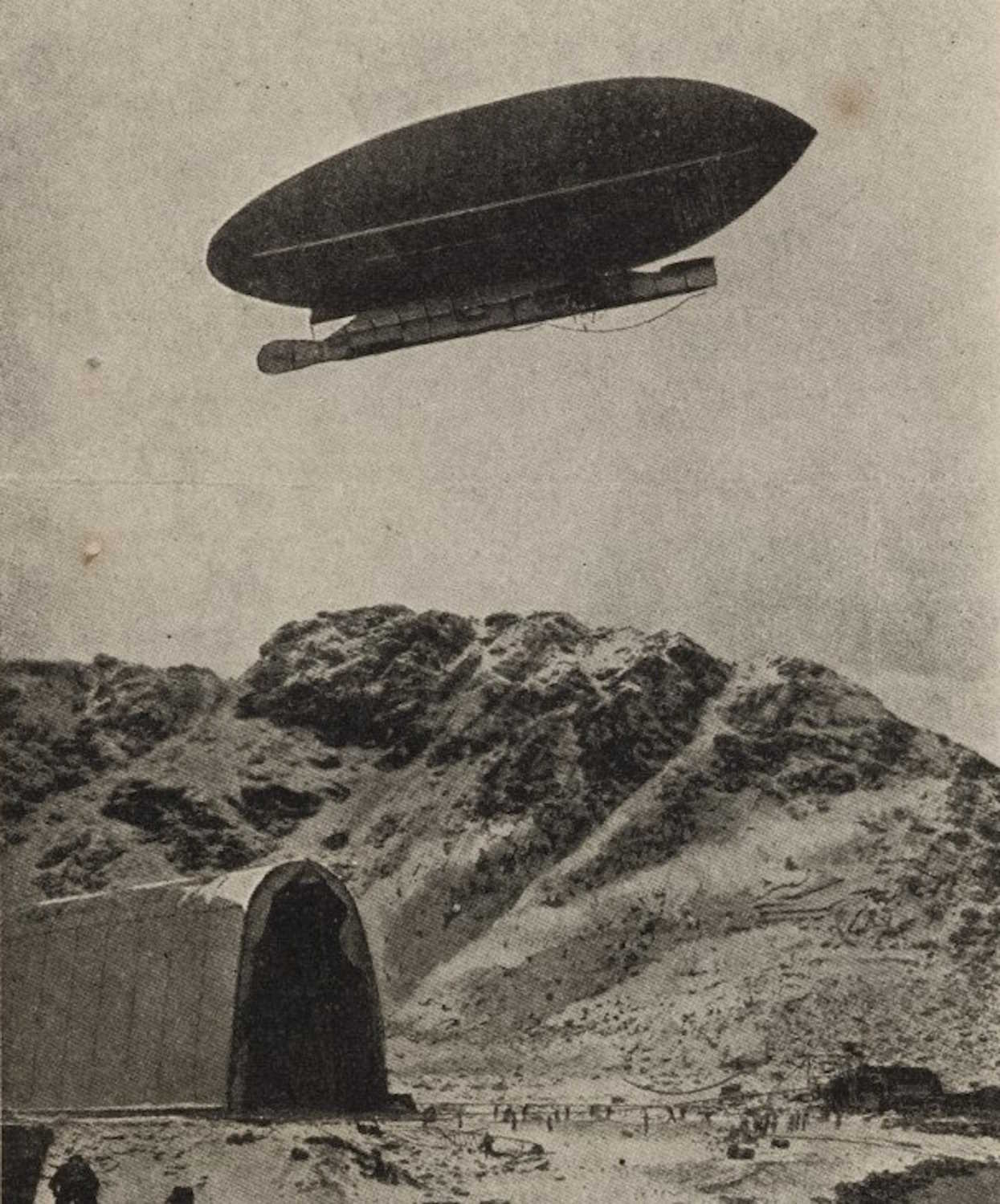 Luftskipet "America" svevende over Virgohamna på Svalbard. Bildet viser et luftskip som svever over et snødekt, fjellandskap. I forgrunnen står en stor, buet hangarlignende bygning med åpen inngang vendt mot kamera. Bak bygningen og i bakgrunnen reiser det seg bratte fjellsider. Under luftskipet kan man skimte små menneskeskikkelser og utstyr på bakken. Hele motivet har et gammelt, kornete preg som tyder på at bildet er historisk.