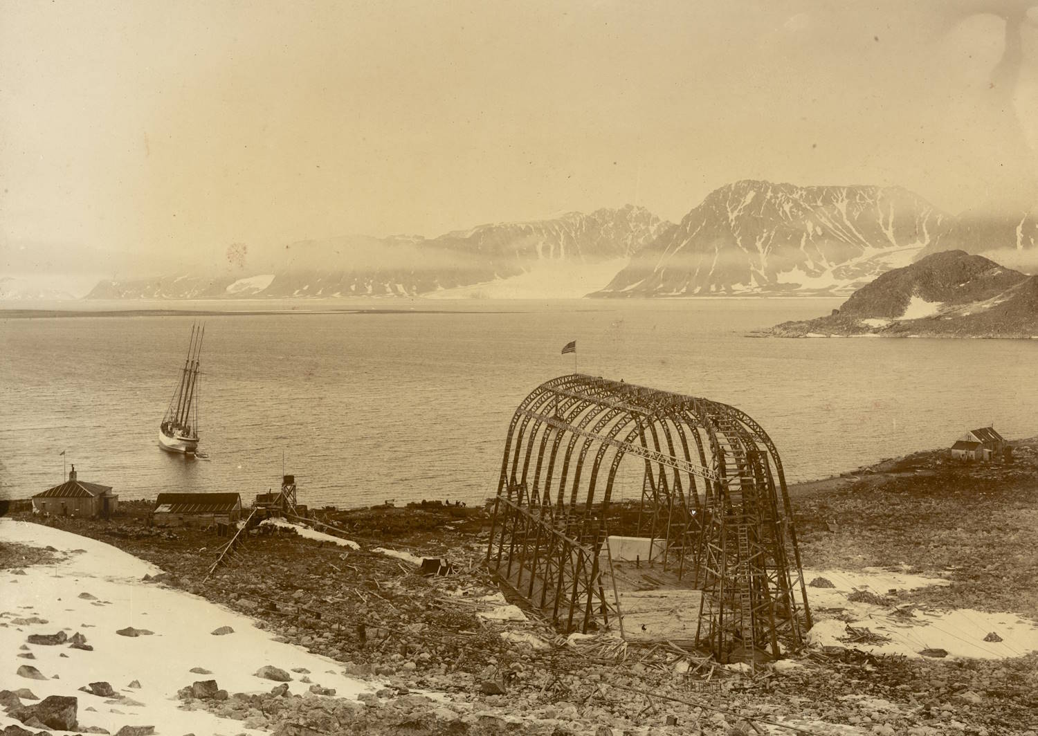 Luftskiphangaren i Virgohamna på Svalbard med motorskonnerten "Artic" liggende i bukta. Bildet viser et karrig kystlandskap med snøflekker i forgrunnen og en stor, buet reisverk‑konstruksjon uten vegger midt i motivet. Konstruksjonen står på et flatt område nær sjøen. Til venstre ligger et seilfartøy for anker, med flere master som peker skrått til siden. Rundt konstruksjonen finnes noen små bygninger og utstyr spredt i terrenget. I bakgrunnen ses en bred fjord omgitt av høye fjell med snø i dalsider og på topper. Lyset er jevnt og gir inntrykk av et gammelt fotografi tatt i arktiske omgivelser.