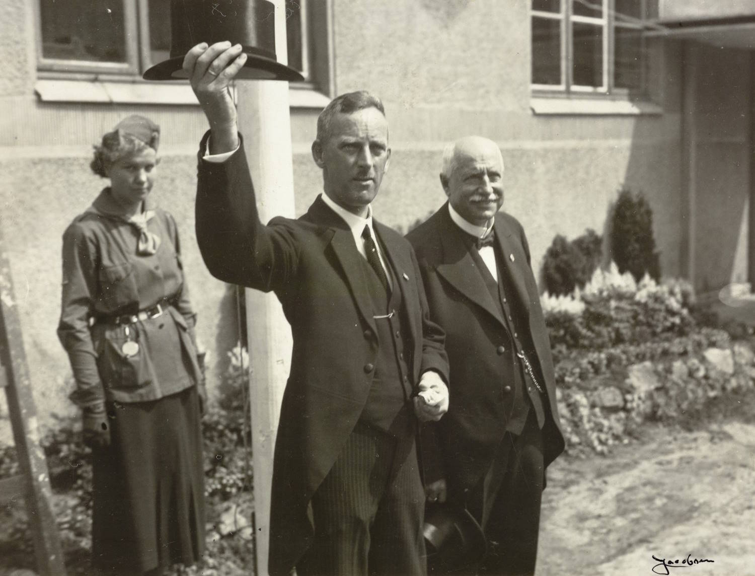 Messesekretær Oscar Spieler og bryggerieier Wilhelm Christiansen under åpningen av varemessen i Arendal 11.07.1936. Foto: Jacobsen. Bildet viser tre personer stående utendørs foran en bygning med lyse murvegger og store vinduer. To menn står i forgrunnen, begge kledd i formelle mørke dresser fra 1930‑tallet. Den ene mannen løfter en høy hatt (flosshatt) i været med strak arm, som om han hilser eller markerer noe. Den andre mannen står tett ved siden av ham og holder en hatt i hånden ned langs siden. Bak dem står en kvinne iført en lang, mørk skjørtedrakt og jakke med belte i livet. Hun har på seg en hatt med brem og står litt tilbaketrukket, vendt mot samme retning som mennene. I bakgrunnen sees et lite oppmurt blomsterbed med lave planter. Det hele fremstår som en formell anledning eller seremoni.