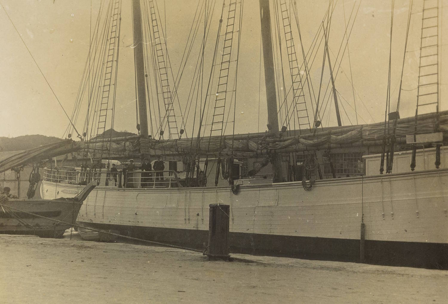 Motorskonnert "Artic" liggende til kai. Bildet viser et stort tremastet fartøy som ligger fortøyd ved en kai. Skipssiden fyller mesteparten av motivet, og mastene strekker seg høyt opp og ut av bildekanten. Langs dekk sees tauverk, stiger og utstyr som henger ned mot skipssiden. Foran skipet ligger en mindre båt inntil skroget. I bakgrunnen skimtes lave åser eller landkonturer. Bildet har et dempet, sepiafarget preg som gir inntrykk av at det er historisk.