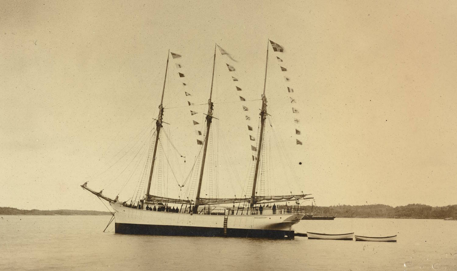 Motorskonnert "Artic" til ankers. Bildet viser et tremastet fartøy som ligger for anker på stille vann. Langs alle tre mastene henger en rekke små vimpler i tauverk som går skrått opp mot mastetoppene. Fartøyet ligger rolig med skroget vendt mot venstre. På høyre side ligger to små robåter fortøyd tett inntil hverandre. I bakgrunnen ses lave, skogkledde øyer eller holmer. Lyset er jevnt og bildet har et gammelt, sepiafarget preg.