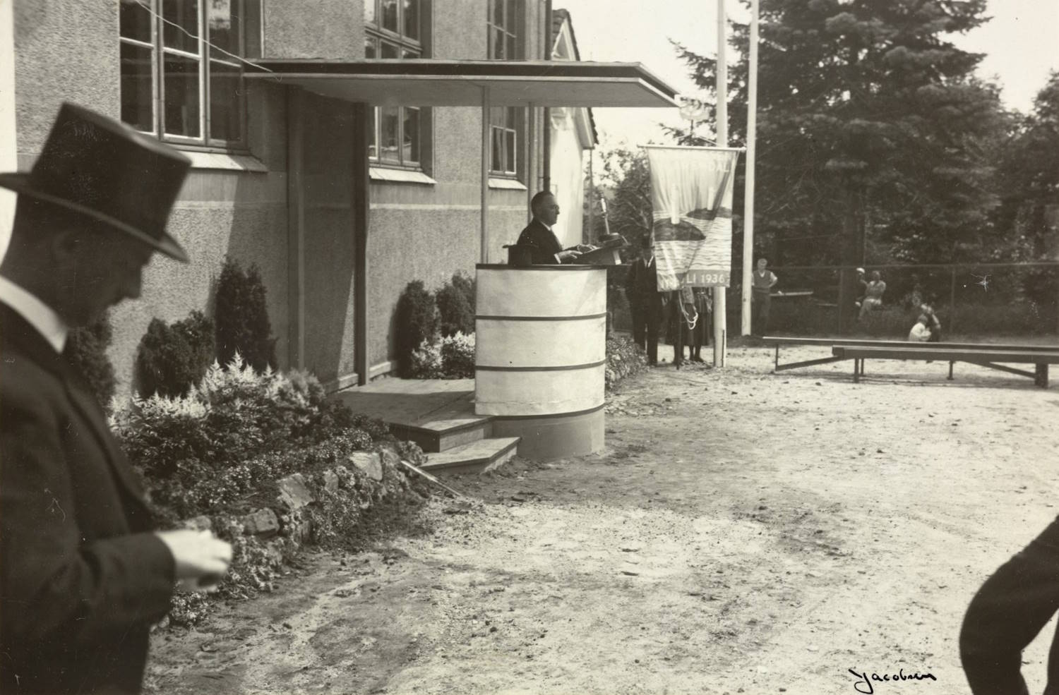 Ordfører Niels Sødring Barth holder tale ved åpningen av varemessen i Arendal 11.07.1936. Foto: Jacobsen. Bildet viser en utendørsscene foran en lys murbygning med store vinduer. Midt i motivet står en talerstol plassert på et lite podium. En mann står bak talerstolen og ser ut til å holde en tale. Talerstolen har et sylinderformet design med horisontale bånd. Til venstre i bildet står en mann i mørk dress og høy hatt. Han er vendt delvis bort fra kamera og ser ned på noe han holder i hendene, muligens et papir eller en liten gjenstand. Bak taleren står et banner eller flagg på en stang, delvis skjult av vegetasjon. I bakgrunnen, til høyre, står flere mennesker, noen bak et gjerde og noen ved en benk. Det er trær og busker rundt området, som gir inntrykk av en formell utendørs seremoni. Foran bygningen ligger et opparbeidet blomsterbed med lav beplantning.
