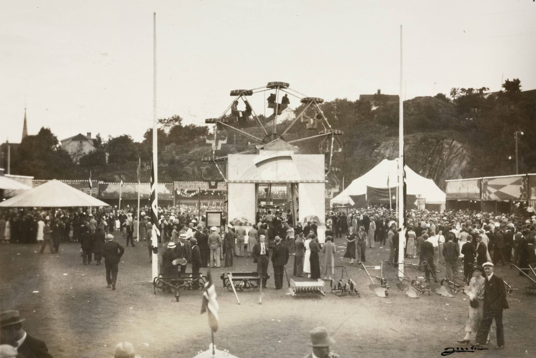 Pariserhjul i fornøyelsesavdelingen på Idrettsplassen under varemessen i Arendal i 1936. Foto: Jacobsen. Bildet viser et livlig messeområde utendørs, fylt med mennesker. Midt i motivet står et stort pariserhjul som rager over resten av området. Hjulet har flere gondoler og er montert på en høy, trekantet konstruksjon. Under pariserhjulet står en portal- eller inngangslignende struktur med tak og åpning på begge sider. Rundt attraksjonen er det mange mennesker som står, går eller betrakter aktivitetene. De fleste er kledd i typiske 1930‑tallsklær: mørke dresser, hatter, kjoler og lange frakker. På begge sider av pariserhjulet står større telt—et kvadratisk telt til venstre og et større, lyst sirkustelt til høyre. Flere flaggstenger står på området, men flaggene er knapt synlige på grunn av bildevinkelen. I forgrunnen er det åpent gress- eller jorddekke, og noen landbruksredskaper som river og harver er stilt opp på bakken. Bak i bildet ligger en liten skogkledd høyde og enkelte hus. Stemningen er folkerik og livlig, som på en stor utendørs messe eller tivoliavdeling.