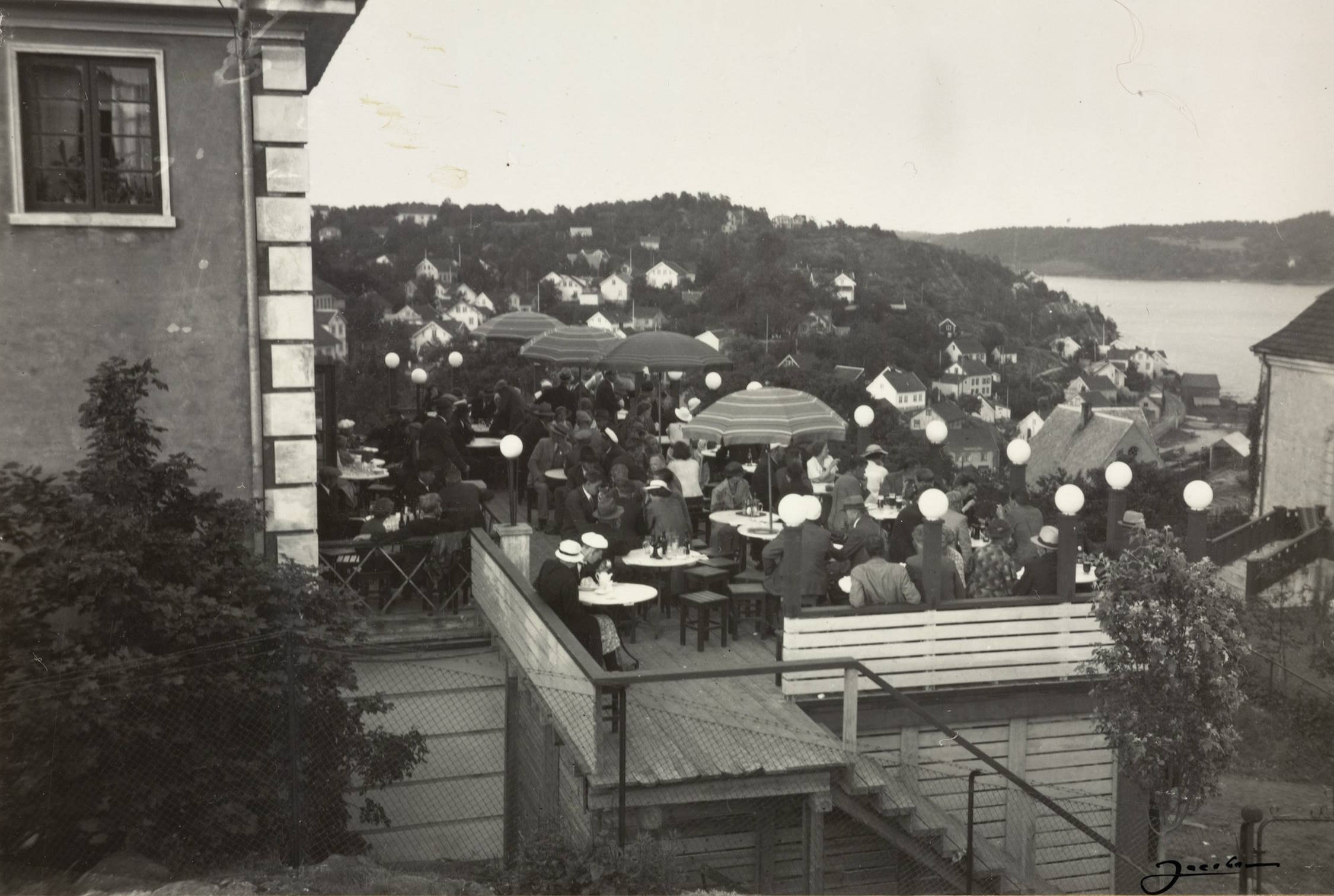 Restauranten ved Sangerhallen under varemessen i Arendal i 1936. Foto: Jacobsen. Bildet viser en utendørs restaurantterrasse med mange mennesker samlet rundt bord. Terrassen består av flere nivåer bygget i tre, forbundet med trapper og rekkverk. Rundt bordene sitter og står personer kledd i typiske 1930‑tallsklær, blant annet dresser, hatter, kjoler og kåper. Flere store parasoller med stripete mønster står spredt mellom bordene. Små lamper på stolper, som ser ut som hvite lys-globuser, er plassert rundt kanten av terrassen og mellom sittegruppene, noe som gir et festpreget inntrykk. Terrassen ligger høyt i terrenget, og i bakgrunnen breier et stort boligområde seg utover en skrånende åsside med mange hvite trehus. Lenger bak ses vann og en åpen kystlinje. Terrassen er delvis omkranset av beplantning og gjerder i tre og netting. Til venstre i bildet står en større bygning med murvegg og vindu, tett inntil terrassen.