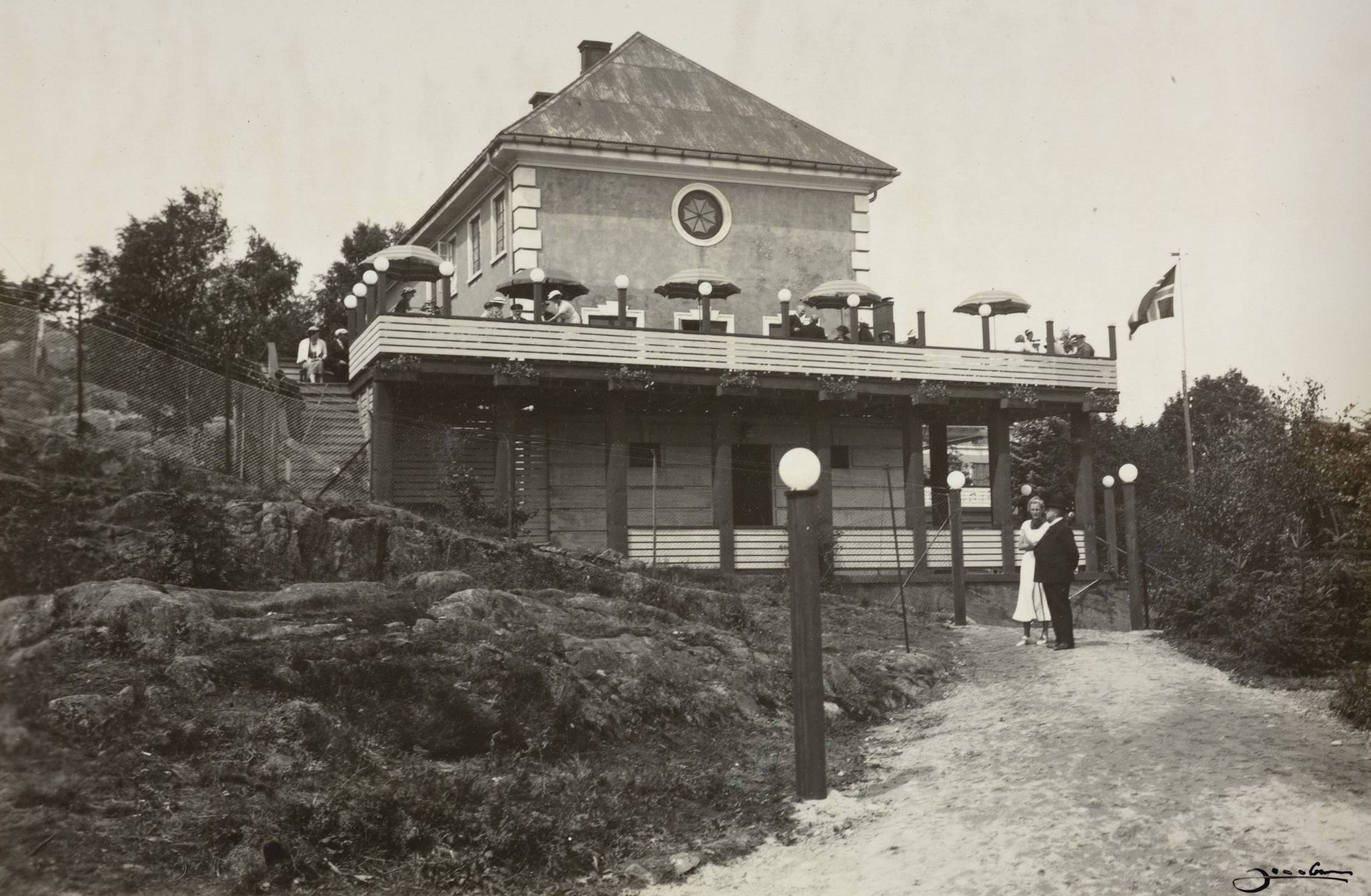 Sangerhallen under varemessen i Arendal i 1936. Foto: Jacobsen. Bildet viser en stor, toetasjes bygning med lys murfasade og høyt, valmet tak. Bygningen ligger i et kupert terreng med bergknauser og stier rundt. Den øverste etasjen har en stor veranda eller terrasse som strekker seg langs hele langsiden. På terrassen sitter og står mennesker ved små bord, flere av dem under stripete parasoller. Terrassen bæres av stolper og bindingsverkskonstruksjon, som delvis skjuler underetasjen. Foran bygningen fører en bred sti, omgitt av gress og busker, opp mot et gjerde og noen trapper som gir adkomst til terrassen. Langs stien står lamper på stolper — runde, hvite glasskuler som ser ut som elektriske utelys. Til høyre i bildet står et flagg på en flaggstang, flagget er heist og synlig fra siden. Terrenget rundt består av nakne svaberg, små trær og tett vegetasjon. Stemningen er rolig og ordnet, og motivet viser et utendørs serveringssted eller samlingsplass på en høydetopp, sannsynligvis i forbindelse med et større arrangement.