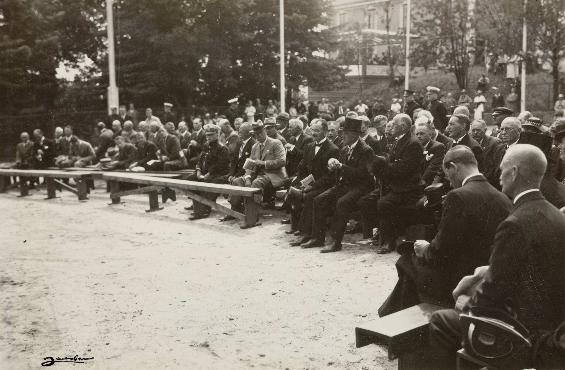 Tilhørere på festplassen ved Barbu skole under åpningen av varemessen i Arendal 11.07.1936. Foto: Jacobsen. Bildet viser en stor forsamling mennesker som sitter og står på en åpen plass utendørs. Publikum sitter på lange trebenker som er satt opp i rekker på et område med hardpakket jord. Benkene er plassert slik at alle vender mot venstre side av bildet, der det trolig foregår en tale eller seremoni som ikke er synlig i rammen.