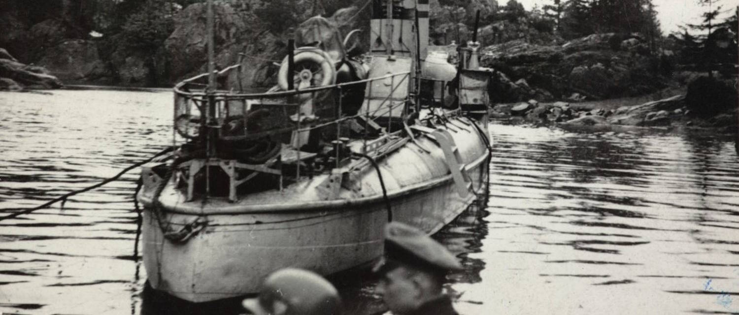Torpedobåten "Grib" ved Lyngør 20. april 1940. Bildet viser en liten militær båt som ligger stille i rolig vann omgitt av svaberg og furutrær. Båten er av eldre type, og skroget virker slitt og skadet. Den har et smalt, avlangt skrog med rekkverk rundt fordekket. Flere kabler og tau henger uordentlig langs sidene. Midt på båten står en høy mast eller stang, og bak den et tårn- eller skorsteinslignende overbygg. På dekk ligger utstyr og deler som ser ustrukturert eller ødelagt ut, blant annet et hjul eller ringformet objekt. I forgrunnen, nederst i bildet, sees hodene til to personer iført militærhjelmer og uniformsluer. Disse er delvis ute av fokus og står nær fotografen. Bak båten ligger kystlandskapet med lave knauser og spredte trær under en lys og overskyet himmel. Stemningen er rolig, men bildet gir et inntrykk av havari eller militær aktivitet.