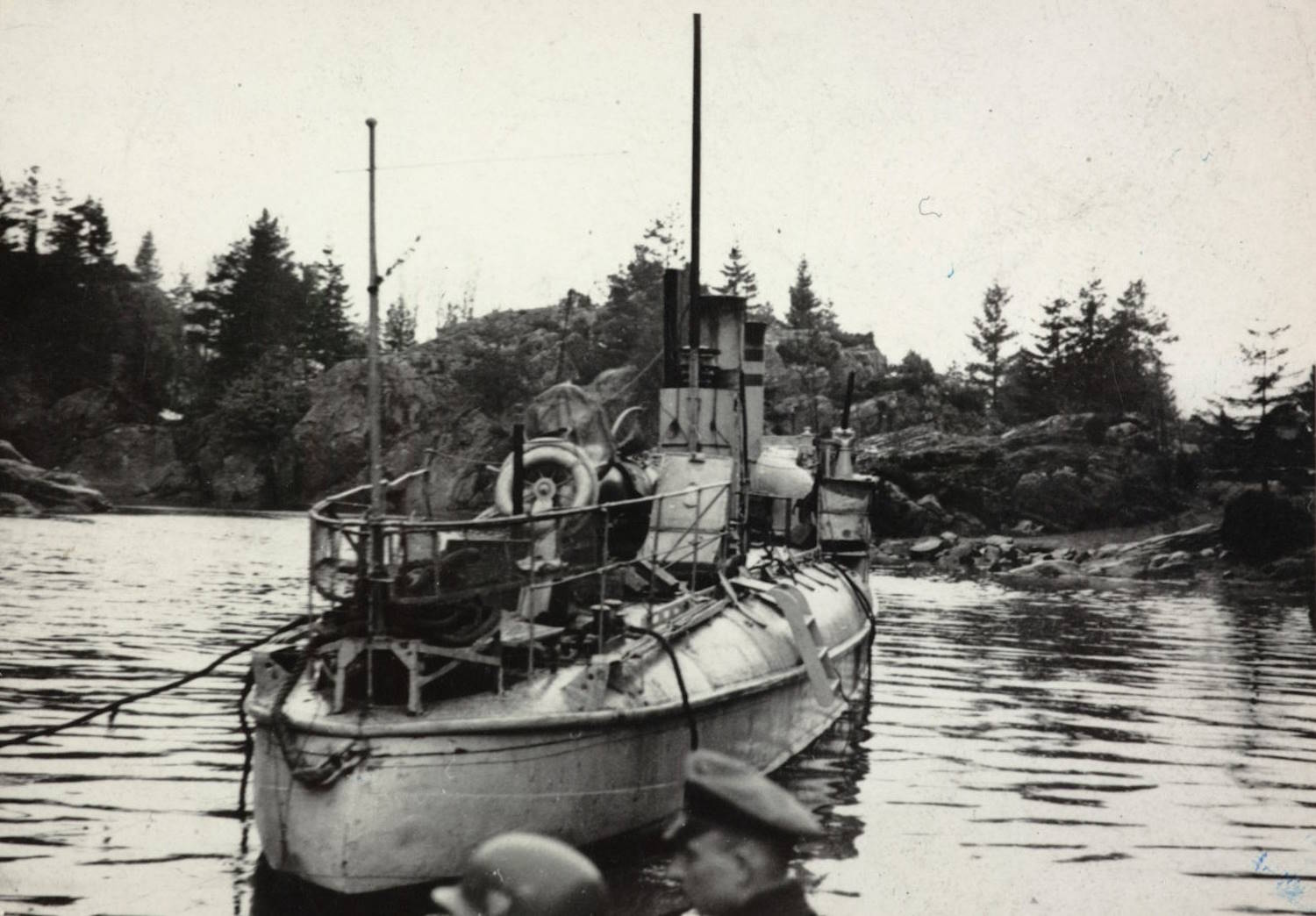 Torpedobåten "Grib" ved Lyngør 20. april 1940. Bildet viser en liten militær båt som ligger stille i rolig vann omgitt av svaberg og furutrær. Båten er av eldre type, og skroget virker slitt og skadet. Den har et smalt, avlangt skrog med rekkverk rundt fordekket. Flere kabler og tau henger uordentlig langs sidene. Midt på båten står en høy mast eller stang, og bak den et tårn- eller skorsteinslignende overbygg. På dekk ligger utstyr og deler som ser ustrukturert eller ødelagt ut, blant annet et hjul eller ringformet objekt. I forgrunnen, nederst i bildet, sees hodene til to personer iført militærhjelmer og uniformsluer. Disse er delvis ute av fokus og står nær fotografen. Bak båten ligger kystlandskapet med lave knauser og spredte trær under en lys og overskyet himmel. Stemningen er rolig, men bildet gir et inntrykk av havari eller militær aktivitet.