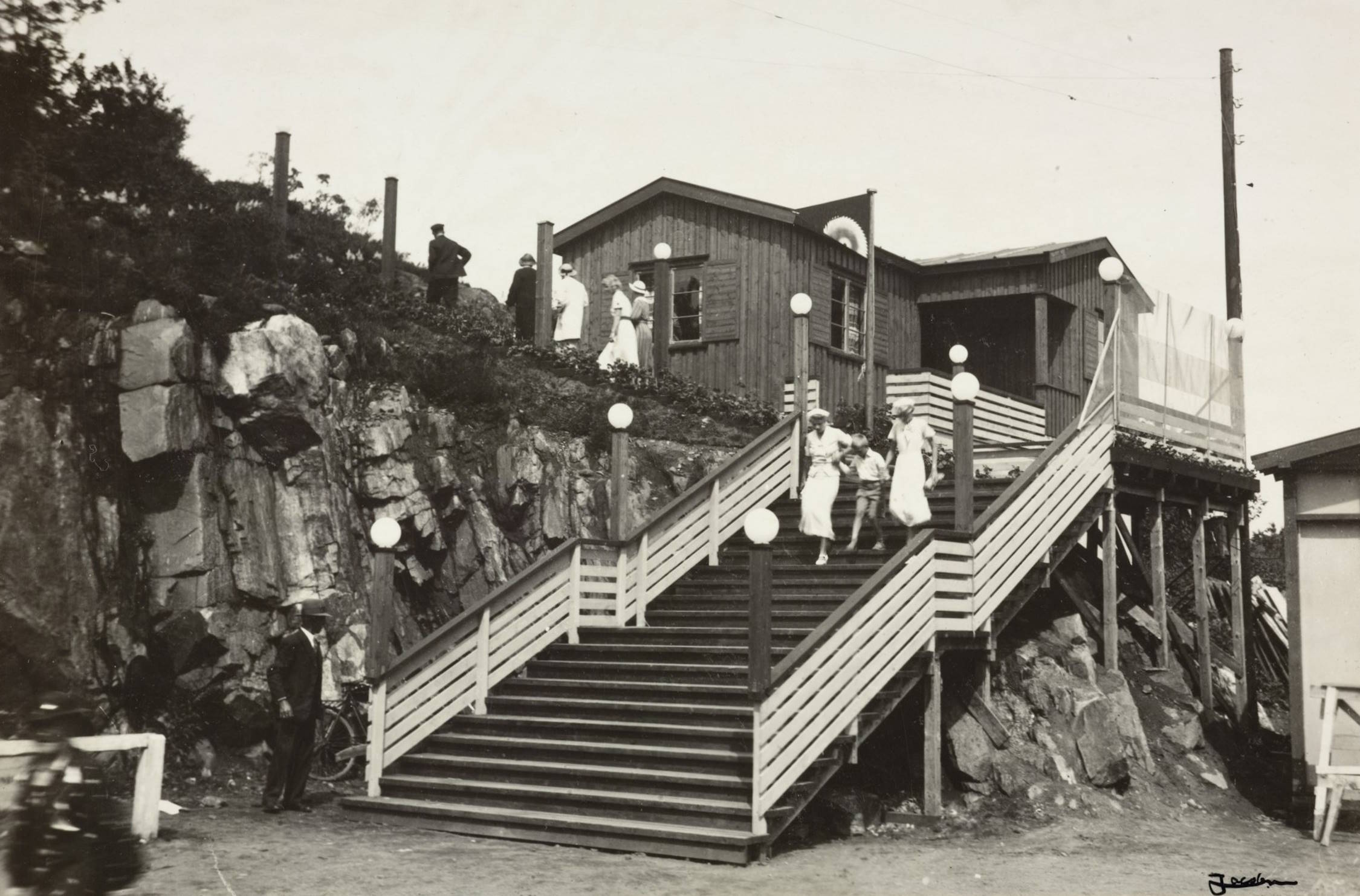 Trapp fra Idrettsplassen på vei mot Sangerhallen under varemessen i Arendal i 1936. Foto: Jacobsen. Bildet viser en utendørstrapp som fører opp en bratt fjellskråning mot en trebygning øverst i bildet. Trappen er bred, har rekkverk på begge sider og er bygget av tre. Langs rekkverket står flere lyktestolper med runde, hvite kupler som gir et inntrykk av belysning langs hele trappeløpet. Øverst på trappen står flere personer kledd i lyse 1930‑tallsklær. To av dem går nedover trappen, mens andre står på flaten ved toppen. De lyse kjolene og hattene gir scenen et sommerlig preg. Til venstre i bildet går fjellet rett opp i ujevne bergflater med noe vegetasjon. Nederst ved trappen står en mann i mørk dress og hatt, vendt delvis bort fra trappen. Trebygningen på toppen har en enkel, funksjonell utforming, med vinduer og en liten terrasse eller platting foran inngangen. Til høyre for bygningen ser man en forhøyning med gjerde og et stort lerret eller skilt festet på veggen. Hele scenen fremstår som en del av et større arrangement, der trappen fungerer som hovedadkomst mellom Idrettsplassen og bygningen oppe i høyden.