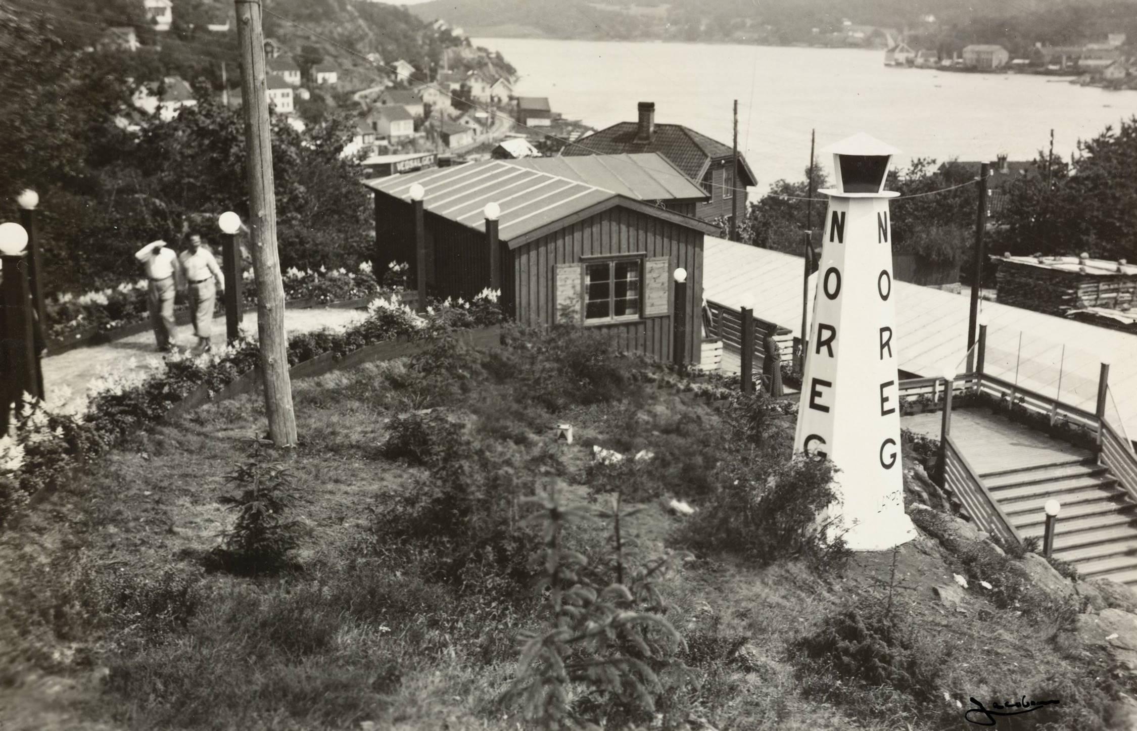 Trapp og sti mellom Idrettsplassen og Sangerhallen under varemessen i Arendal i 1936. Foto: Jacobsen. Bildet viser et kupert uteområde med en grussti som slynger seg oppover en skråning. Vegetasjonen består av gress, lave busker og enkelte små trær. Til høyre i bildet står en høy, hvit trekonstruksjon som ligner en dekorativ varde eller et tårn. Den er formet som en smal, konisk søyle med et lite tak på toppen. Langs siden står teksten “NOREG” malt i store, mørke bokstaver. Bak denne konstruksjonen går en bred tretrapp eller rampe oppover mot en trebygning. Bygningen er liten, mørk og enkel i formen, med skrått tak og ett vindu vendt mot kamera. Rundt bygningen står flere hvite kuleformede lamper på stolper, som går igjen langs stier og trapper i området. Til venstre for bygningen står to personer kledd i lyse klær og hatter, på en sti som følger terrenget. Lenger i bakgrunnen ser man et utsyn over et større boligområde med mange små trehus som ligger spredt utover skrånende terreng. Bak husene ligger en bred vannflate, trolig en fjord eller bukt, og åser strekker seg opp på motsatt side. Hele motivet gir inntrykk av en utsiktsplass eller et område knyttet til et større arrangement, med tydelig tilrettelegging for publikum gjennom trapper, stier og belysning.
