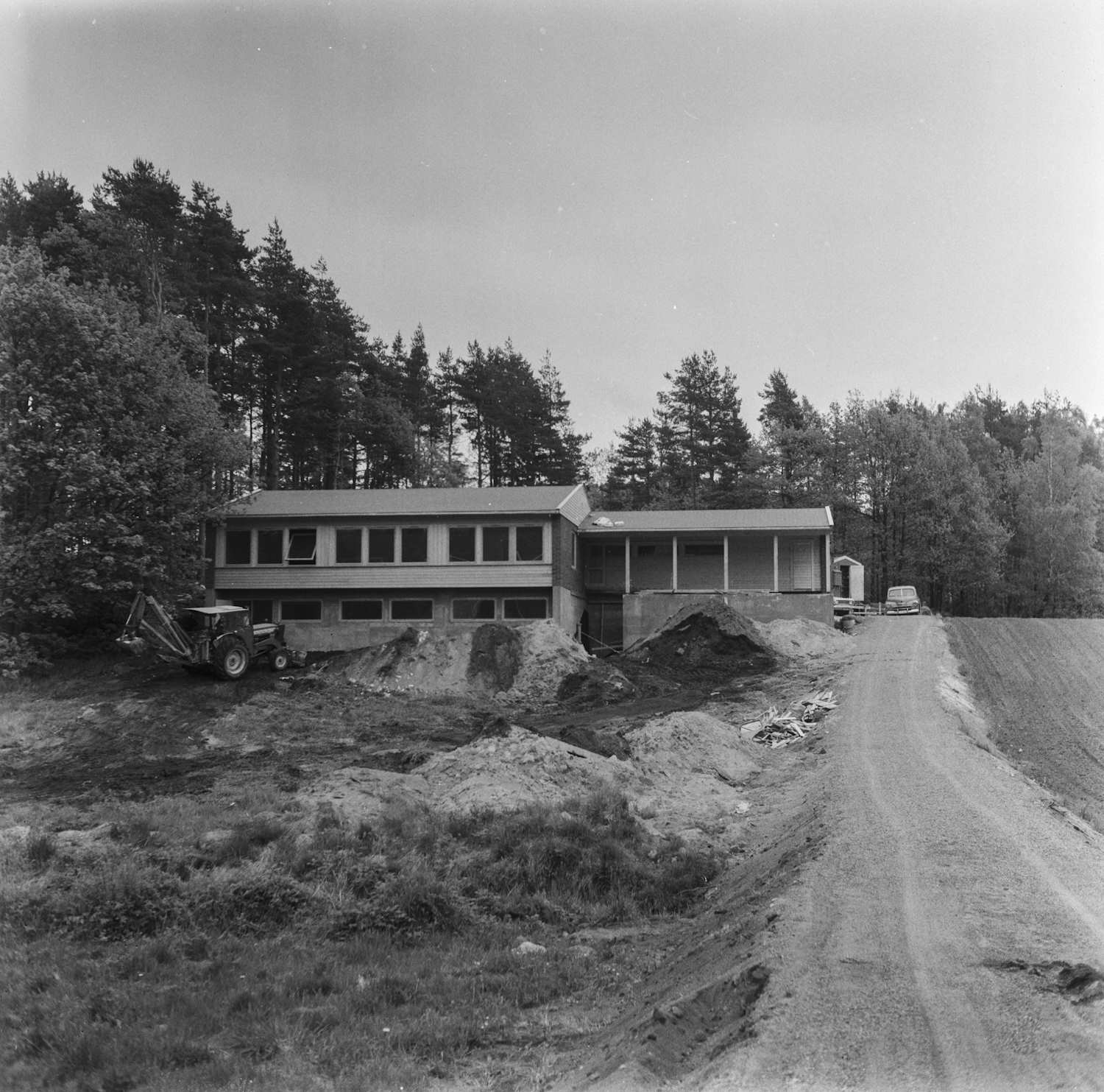 Ellengård daghjem og skole rett før åpningen i august 1966. Foto: Per Skjeggestad. Bildet er et sort-hvitt fotografi som viser Ellengård daghjem og skole rett før åpningen i august 1966. I midten av bildet står en lang, lav bygning i to etasjer med flatt tak og store vinduer. Bygningen ser nyoppført ut, og foran den ligger hauger med jord og byggematerialer. Til venstre foran bygningen står en traktor med skuff, parkert på ujevnt terreng. Området rundt fremstår som en byggeplass, med bar jord og spor etter anleggsmaskiner. Til høyre går en grusvei forbi bygningen, og lenger bak på veien står en bil parkert. Bak bygningen og langs venstresiden av bildet er det tett skog med høye trær. Himlingen er lys og uten markerte skyer. Det er ingen mennesker synlige i bildet. Stemningen er rolig og dokumenterende, med fokus på bygningen og omgivelsene før ferdigstillelse.