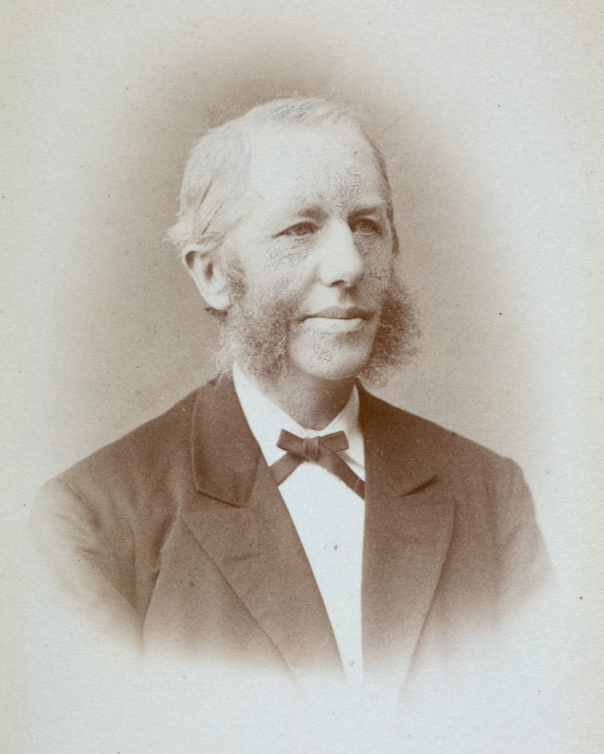 Ernst Hadeler Ziesler. Bildet er et gammelt portrettfotografi i lyse, sepiafargede toner med myk, utydelig kant rundt motivet. Bakgrunnen er ensfarget og lys, uten synlige detaljer. En person er avbildet fra brystet og opp, vendt rett mot kamera. Vedkommende har lyst hår som er kjemmet bakover, og et øre er synlig på venstre side. Personen er kledd i en mørk dressjakke over en lys skjorte, med en liten sløyfe i halsen. Fotografiet har et rolig og formelt preg, typisk for eldre portrettfotografi, med jevn belysning og et tidløst uttrykk.