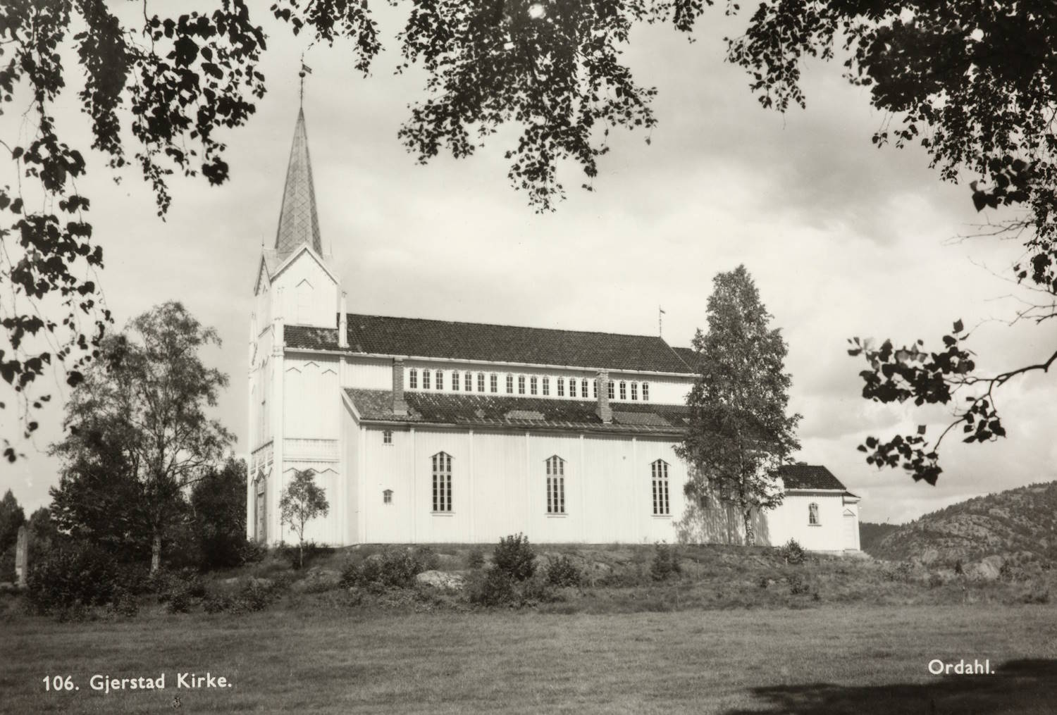 Bildet er et svart-hvitt fotografi av Gjerstad kirke, sett fra siden i et åpent landskap. Kirken er en stor, hvitmalt trekirke med rektangulær grunnform og et høyt, smalt tårn med spiss avslutning plassert i venstre ende av bygningen. Tårnet har en værhane på toppen. Langs kirkens langside er det flere høye, smale vinduer jevnt fordelt. Taket er mørkt og skrått, og midt på takflaten sees en forhøyet del med små vinduer. Til høyre for hovedbygget er et lavere tilbygg. Kirken står på en svakt forhøyet gressbakke. I forgrunnen er det åpent gressområde. Rundt kirken står spredte løvtrær, og greiner med blader rammer inn bildet øverst. I bakgrunnen anes lave åser eller skogkledd terreng. Stemningen er rolig og tidløs, og bildet fremstår som et eldre dokumentasjonsfoto. Nederst i bildet står teksten «106. Gjerstad Kirke.» samt fotografnavnet «Ordahl».