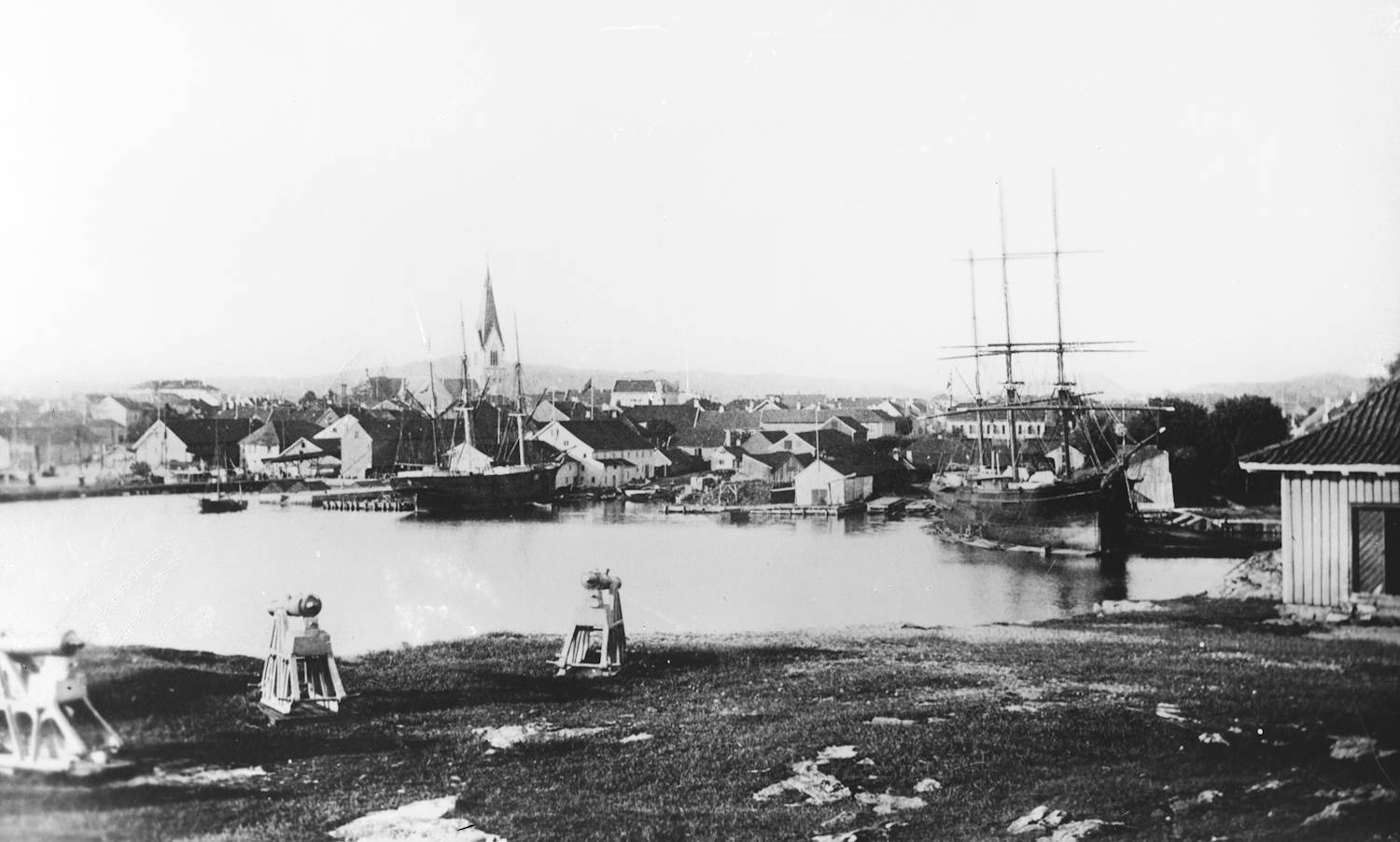 Kristiansand 1891. Fotograf: Lindstrøm. Historisk sort-hvitt foto av havnen med mange lave trehus langs vannet. I bakgrunnen et kirkespir. I havnebassenget ligger to store seilskip med høye master og en mindre seilbåt. Foran vannet er en gresskledd slette med to kanoner og en enkel trebygning til høyre.