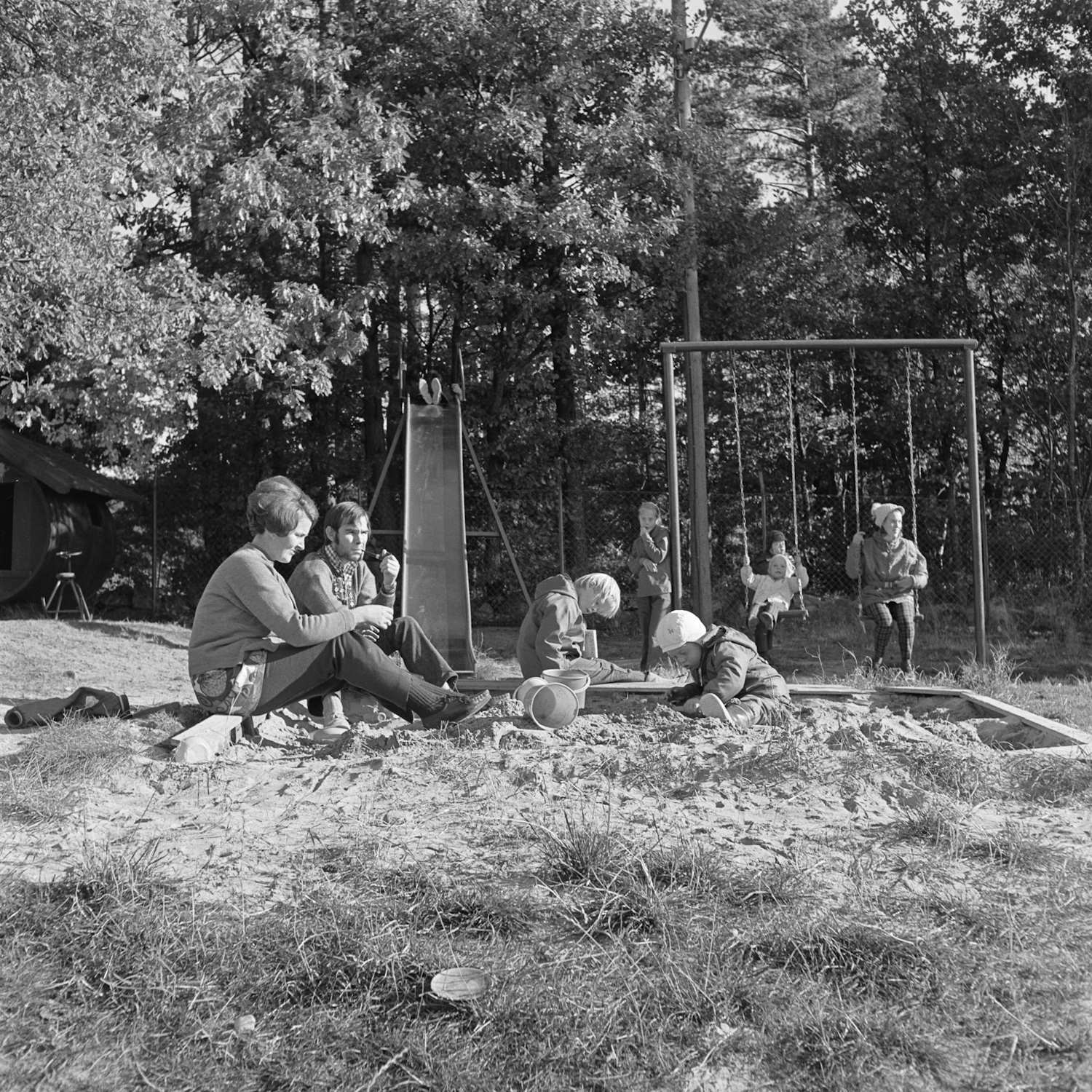 Uteaktiviteter ved Ellengård i oktober 1971. Bildet er et svart-hvitt fotografi tatt utendørs i et skogkledd område. I forgrunnen sitter flere barn i en sandkasse og leker med bøtter og spader. Barna har på seg yttertøy som jakker, bukser og luer, noe som tyder på kjølig høstvær. Til venstre for sandkassen sitter to voksne på bakken. Den ene er en dame som sitter vendt mot barna og følger med på leken. Ved siden av henne sitter en mann som holder en pipe. Begge har på seg ytterklær og fremstår som tilsynspersoner. Midt i bildet står en lav sklie, og til høyre er det et huskestativ. Flere barn oppholder seg der, noen sitter på huskene mens andre står i nærheten. Bakgrunnen består av høye løvtrær med tett bladverk. Området er preget av sand og gress, og helhetsinntrykket er et rolig og hverdagslig uteområde, som for eksempel en barnehage eller et institusjonsmiljø.