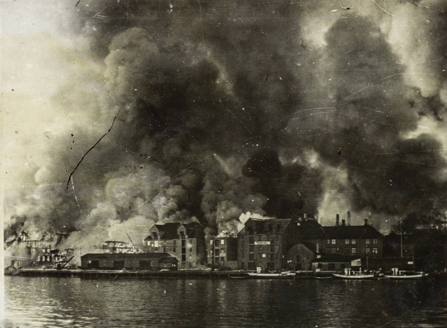 Kristiansund i flammer 1940. Bildet er et sort-hvitt fotografi som viser en tettbygd kystbebyggelse langs en vannflate. I bakgrunnen reiser det seg store, mørke røykskyer som dekker store deler av himmelen. Røyken ser tett og urolig ut og sprer seg horisontalt over byen. Langs vannkanten står flere bygninger i mur og tre, noen med synlige tak og fasader. Flere av bygningene er delvis skjult av røyk, og enkelte steder kan det skimtes lysere partier som kan tyde på brann eller kraftig varmeutvikling. Bygningene ligger tett i rekker, med varierende høyde og takformer. I forgrunnen ligger rolig vann, med speilinger av bygningene og røyken. Det er små båter eller fartøy fortøyd langs kaien på høyre side av bildet. Det finnes ingen tydelig synlige personer i bildet. Helhetsinntrykket er en urban kystby rammet av en omfattende brann, med kraftig røykutvikling som dominerer motivet og gir bildet et dramatisk og alvorlig preg.