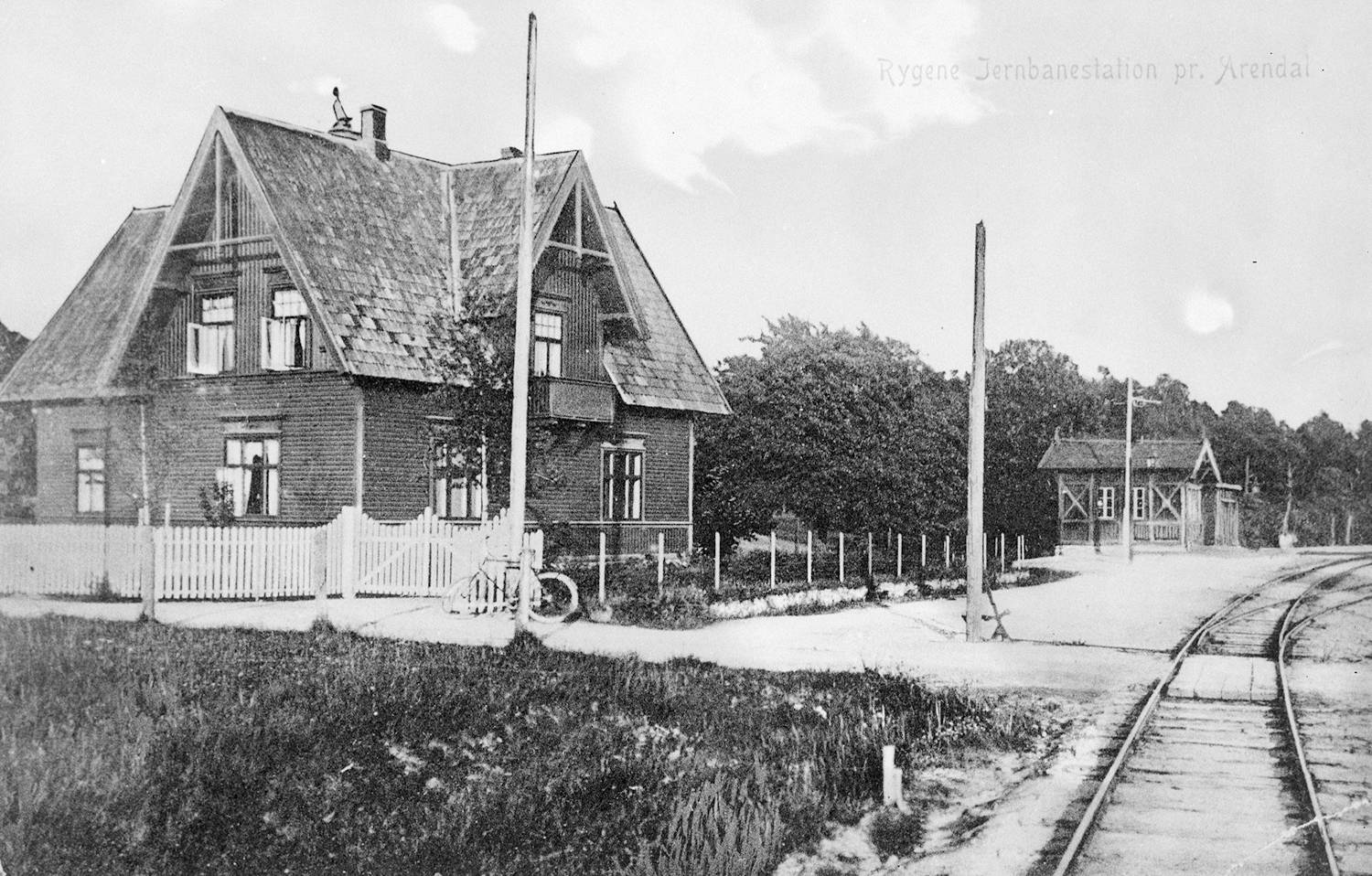 Arbos hus på Fossum i Øyestad. Bildet er et svart-hvitt fotografi av et jernbaneområde i landlige omgivelser. I forgrunnen til venstre står et toetasjes trehus med bratt saltak og flere vinduer. Huset har et lite inngjerdet område med hvitt stakittgjerde. Ved gjerdet, nær inngangen, står en sykkel lent mot gjerdet. Foran og til høyre i bildet går jernbanespor som buer svakt innover i bildet. Langs sporet står flere høye stolper, trolig til strøm eller signaler. Bak huset og langs sporet er det lave gjerder og åpne grus- og gressflater. I bakgrunnen, til høyre, ses en mindre bygning med lignende arkitektonisk stil, plassert ved siden av jernbanen. Bak denne bygningen er det tett vegetasjon med trær som danner en grønn bakgrunn, selv om fargene fremstår i gråtoner. Øverst til høyre i bildet er det trykt tekst som leses: «Rygene Jernbanestasjon pr. Arendal». Bildet fremstår som et historisk fotografi uten synlige mennesker, tatt i dagslys med jevn belysning.