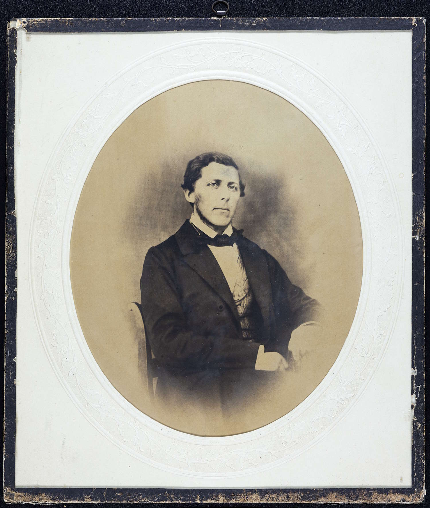 Axel Christian Smith Arbo. Bildet er et eldre portrettfotografi presentert i et rektangulært monteringskort med mørk ytterkant og en lys, dekorert oval ramme rundt selve fotografiet. Midt i bildet, innenfor den ovale rammen, sitter en person på en stol i halvprofil, vendt svakt mot høyre. Personen har på seg mørk jakke, lys skjorte og vest, samt et mørkt halstørkle eller slips. Hendene er samlet foran kroppen. Kroppsholdningen er oppreist og formell. Bakgrunnen i portrettet er jevnt tonet i varme brun- og sepiafarger, uten tydelige detaljer, noe som gir et mykt og nøytralt bakteppe. Bildet fremstår som et studioportrett fra 1800-tallet, med rolig komposisjon og tydelig fokus på personen.