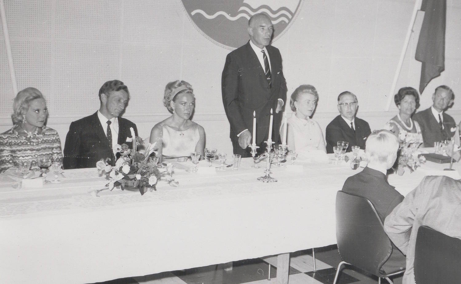 Direktør ved Akers mek. Verksted Onar Onarheim takker for oppdraget under festmiddagen 11. juli 1968. Sonja Henie Onstad sittende helt til venstre. Bildet er et svart-hvitt fotografi fra en formell middag i et innendørs lokale. En lang, hvitdekket festbordrekke strekker seg horisontalt gjennom bildet. Rundt bordet sitter flere personer tett ved siden av hverandre. Midt i bildet står én person oppreist bak bordet, kledd i mørk dress og slips. De øvrige personene sitter, også iført formelle klær som dresser, kjoler og smykker. På bordet står blomsteroppsatser, lysestaker med tente lys, glass, tallerkener og servise. Borddekkingen fremstår symmetrisk og gjennomført. I bakgrunnen ses en lys vegg med et rundt emblem eller dekorasjon øverst, samt et flagg plassert på høyre side av bildet. I forgrunnen, nederst til høyre, ses ryggen av en person som sitter ved et bord vendt mot festbordet. Helhetsinntrykket er en høytidelig og seremoniell sammenkomst,