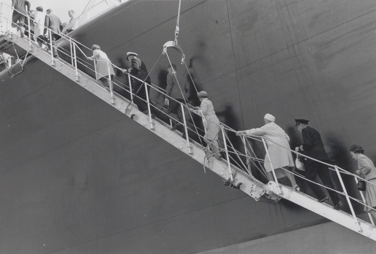 Gjestene går opp gangveien under skipsdåpen av M/T "Credo" 11. juli 1968. Bildet er et svart-hvitt fotografi tatt ved siden av et stort skipsskrog. Skroget fyller nesten hele bakgrunnen og fremstår som en høy, mørk og jevn flate av stål. I forgrunnen går en gruppe mennesker opp en smal, skrå gangvei som er festet til skipets side. Gangveien har rekkverk på begge sider og strekker seg diagonalt fra nedre høyre hjørne av bildet og opp mot øvre venstre. Personene beveger seg i samme retning, oppover mot skipet. Menneskene er kledd i yttertøy som frakker og hatter, noe som tyder på en formell anledning. Noen bærer lyse klær, andre mørkere. Enkelte holder i rekkverket mens de går. Det er relativt tett mellom personene på gangveien. Det er ingen synlig tekst i selve bildet.