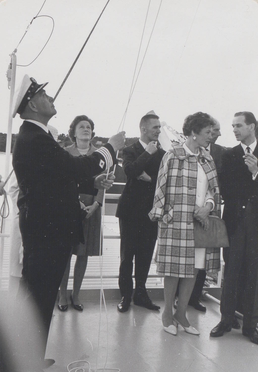 Kaptein Martin Olsen foretar flaggskiftet under skipsdåpen til M/T "Credo" 11. juli 1968. Bildet er et svart-hvitt fotografi tatt utendørs på dekk av et skip. Dekket er lyst og åpent, og bakgrunnen viser en lav horisontlinje med land eller trær i det fjerne. I venstre del av bildet står en person i mørk uniform med lue. Personen holder et tau eller en line som går oppover til en flaggstang eller vaier. Tauet er stramt og tydelig i forgrunnen. Midt i bildet står flere personer samlet i formelle klær. Noen har mørke dresser, andre har lyse kåper eller drakter. Én person bærer en rutemønstret kåpe og holder en håndveske. En annen person står litt bak og ser mot tauet og flaggmekanismen. Til høyre i bildet står ytterligere personer tett sammen, også kledd i formelt yttertøy. De er vendt mot handlingen i midten av bildet. Det er synlige tau, vaiere og tekniske detaljer knyttet til skipets rigg. Ingen tekst er synlig i selve bildet. Helhetsinntrykket er en høytidelig handling på skipets dekk, der en formell flaggheising eller et flaggskifte foregår i nærvær av inviterte deltakere.