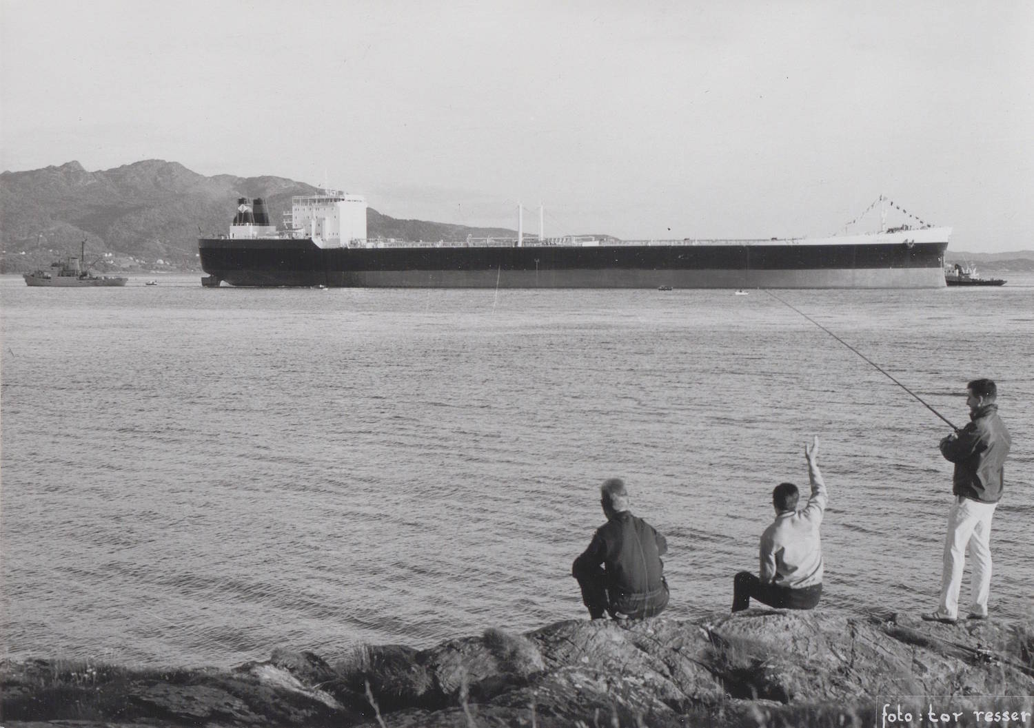 M/T "Credo" på prøvetur 4. eller 5. juli 1968. Foto: Tor Resser. Bildet er et svart-hvitt fotografi tatt utendørs ved sjøen. I midten av bildet ligger et svært langt og lavt skip på vannet, sett i profil. Skipet strekker seg nesten over hele bildebredde. Skroget er mørkt, mens overbygningen akter er lysere og tydelig avtegnet. Det er flere detaljer langs dekket, blant annet master og konstruksjoner som gir inntrykk av et stort tankskip. Mindre fartøy ligger i nærheten av skipet, både foran og bak. I forgrunnen, nederst i bildet, sitter og står tre personer på en steinete strand eller et svaberg. To personer sitter vendt mot sjøen og skipet, mens én person står til høyre og holder en fiskestang som er vinklet utover mot vannet. En av de sittende personene har armen løftet, som om det vinkes mot skipet. Bakgrunnen viser en lav fjellrekke eller kupert landskap langs kysten, som strekker seg horisontalt bak skipet. Himlingen er lys og jevn uten tydelige skyer. Det er synlig tekst nederst til høyre i bildet: «foto: tor tesser». Helhetsinntrykket er et rolig kystmotiv der et stort skip er ute på prøvetur, observert fra land av mennesker i forgrunnen.
