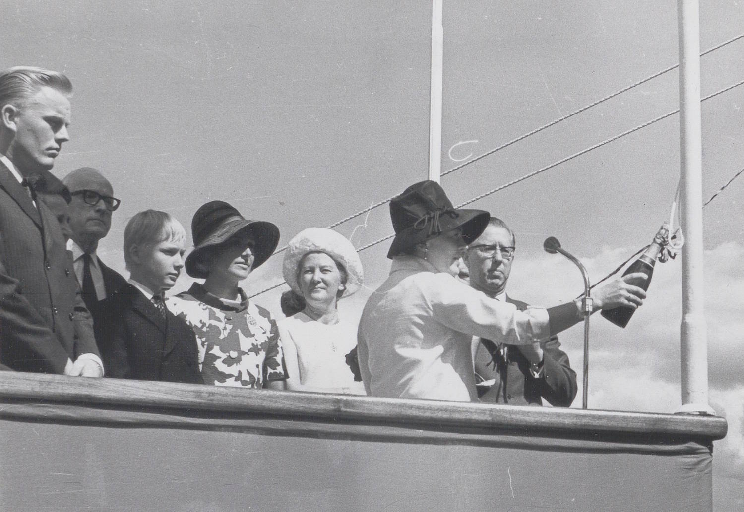 Marit Wangen døper skipet "Credo" 11. juli 1968. Bildet er et svart-hvitt fotografi tatt utendørs på dekk av et skip, under en seremoni. Forgrunnen domineres av en rekke personer som står tett sammen bak et rekkverk langs skipssiden. Til høyre i bildet holder én person en flaske i strak arm, vinklet utover mot skipsiden. Flasken er festet med et bånd eller en snor. Like ved er en mikrofon montert på et stativ, rettet mot personen med flasken. Flere vaiere og tau fra skipets rigg er synlige bak og over personene. De øvrige personene står på rekke og er kledd i formelle klær, som mørke dresser, lyse kåper og hatter. Noen har hodeplagg, andre ikke. Alle vender oppmerksomheten mot handlingen med flasken. Bakgrunnen består av lys himmel med spredte skyer. Ingen tekst er synlig i selve bildet. Helhetsinntrykket er en høytidelig handling på skipets dekk, der en sentral seremoni utføres foran inviterte deltakere.