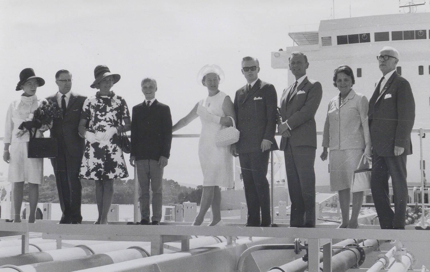 Marlow Wangen med familie og følge på dekk under skipsdåpen til M/T "Credo" 11. juli 1968. Bildet er et svart-hvitt fotografi tatt utendørs på dekk av et stort skip. En gruppe personer står på rekke langs et rekkverk, høyt hevet over dekket. Alle er kledd i formelle klær som dresser, kåper og drakter, og flere bærer hatter. Én person holder en blomsterbukett, mens en annen bærer en håndveske. Personene står tett ved siden av hverandre og vender hovedsakelig fremover og litt mot venstre i bildet. Midt i rekken står en person i lys kjole som har en arm strakt ut mot personen ved siden av, som om det holdes kontakt eller vises vei. I forgrunnen under rekkverket er tekniske detaljer fra skipet synlige, blant annet rør og sylindriske konstruksjoner som løper horisontalt. I bakgrunnen til høyre ruver overbygningen på skipet, med rette linjer, vinduer og dekkstruktur. Bak gruppen ses også en lys himmel. Det er ingen synlig tekst i bildet. Helhetsinntrykket er en formell og høytidelig samling på skipets dekk, der en gruppe inviterte står oppstilt under en seremoni.