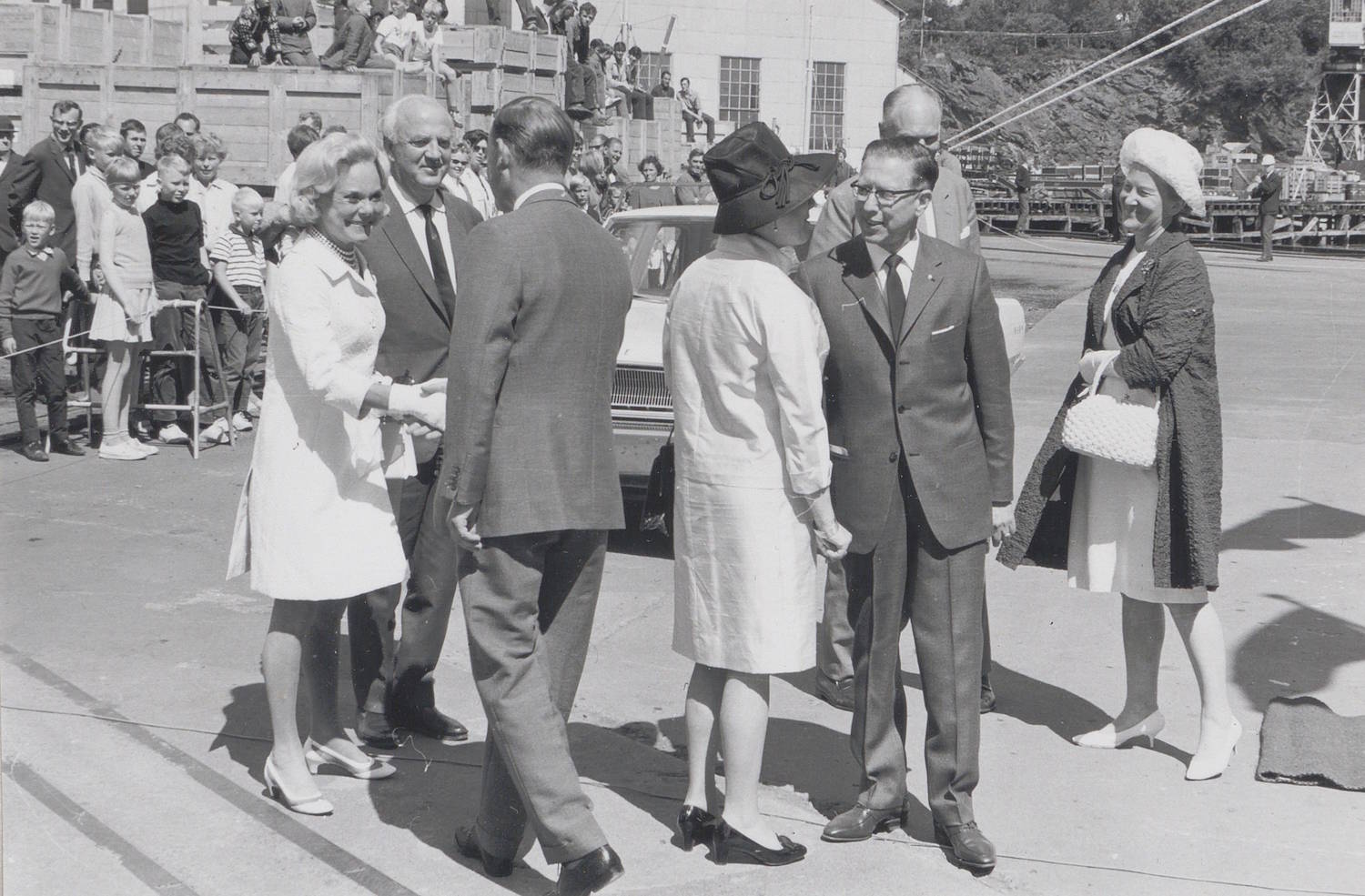 Sonja Henie Onstad og Niels Onstad ønskes velkommen av Marlow Wangen under skipsdåpen til M/T "Credo" 11. juli 1968. Bildet er et svart-hvitt fotografi tatt utendørs på et industrielt område ved sjøen, trolig på eller ved et verft. I forgrunnen står en gruppe personer samlet på bakkenivå, kledd i formelle klær som dresser, kåper og drakter. Flere bærer hatter, og én person holder en håndveske. Personene står i små klynger og vender seg mot hverandre, som om det pågår hilsing eller samtale. Midt i gruppen er to personer vendt mot hverandre med hendene strakt frem, som i en håndhilsning. Bak gruppen står en bil parkert, delvis synlig mellom personene. I bakgrunnen ses en større folkemengde samlet bak et lavt gjerde eller en avsperring. Mange står tett sammen og ser mot gruppen i forgrunnen. Bak folkemengden er det synlige bygninger med rette vegger og vinduer, samt tekniske konstruksjoner, vaiere og kraner som gir inntrykk av et havne- eller verftsområde. Underlaget er asfalt eller betong, og lysforholdene tyder på dagslys med klare skygger. Det er ingen synlig tekst i selve bildet. Helhetsinntrykket er en formell mottakelse eller velkomst i forbindelse med et arrangement, med både inviterte deltakere i forgrunnen og publikum i bakgrunnen.