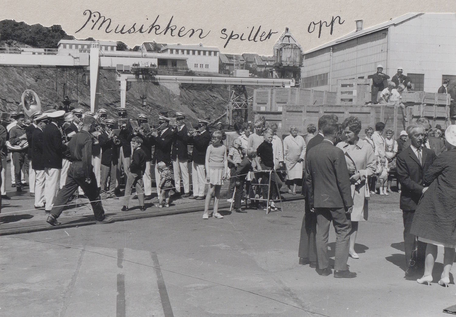 Stord Musikklag spiller under dåpen av M/T "Credo" 11. juli 1968. Bildet er et svart-hvitt fotografi tatt utendørs på et industrielt område ved sjøen, trolig på et skipsverft under et arrangement. I forgrunnen og midten av bildet ses et større folkeansamling samlet på et åpent asfalt- eller betongområde. Til venstre i bildet står et musikkorps i formasjon. Korpsmedlemmene bærer uniformer og holder blåseinstrumenter, blant annet trompeter og en stor tuba som er godt synlig. Bak og rundt korpset står mange tilskuere i ulike aldre, kledd i hverdags- og penklær. Noen står bak en lav avsperring, mens andre står fritt på plassen. Flere barn er synlige blant publikum. I høyre del av bildet står mindre grupper av personer i formelle klær, som dresser og kåper, vendt mot hverandre i samtale. Bak disse gruppene ses industrielle bygninger med rette vegger og vinduer, samt tekniske installasjoner, kraner og vaiere som understreker verftsmiljøet. Øverst i bildet, over bygningene, er det håndskrevet tekst: «Musikken spiller opp». Helhetsinntrykket er en livlig og organisert scene der et musikkorps spiller under en offentlig markering, med mange fremmøtte tilskuere i et tydelig industrielt havnemiljø.