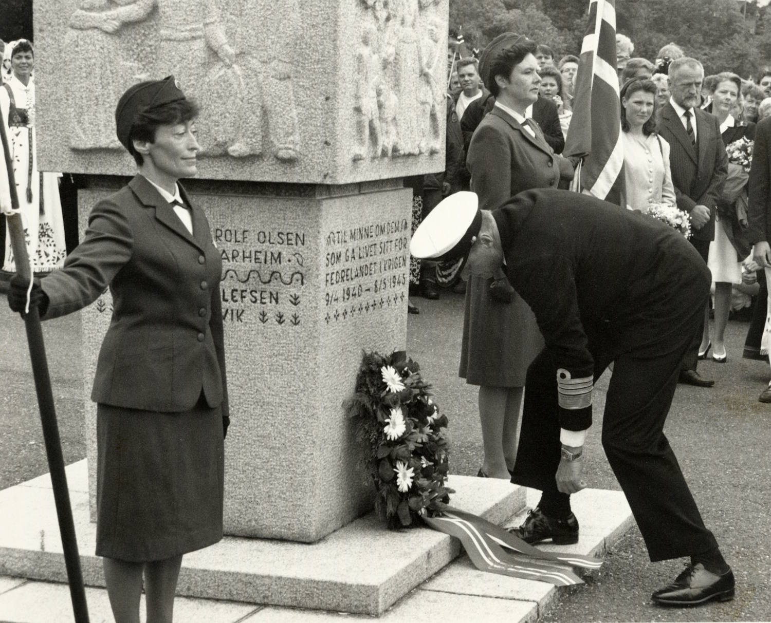 Kong Harald 5. legger ned krans ved kirgsminnestøtten i Grimstad under signingsferden 28.06.1991. Foto: Trolig Erik Holand, Agderposten. vart-hvitt fotografi fra en minneseremoni ved et steinmonument. I forgrunnen står en person i mørk marineuniform med hvit uniformslue bøyd framover ved foten av monumentet for å legge ned en krans. Kransen er dekorert med blomster og bånd som ligger ned langs sokkelen. Til venstre står en person i mørk uniform og holder en fane eller flaggstang. Midt i bildet reiser den store granittstøtten seg med relieff og innhogd tekst. Deler av inskripsjonen er lesbare, blant annet: «Til minne om dem som ga livet sitt for fedrelandet i krigen 1940–1945». Bak monumentet står flere mennesker samlet som tilskuere. Et stort norsk flagg er synlig på høyre side av bildet. I bakgrunnen sees også personer i høytidsklær og bunader. Bildet formidler en høytidelig kransenedleggelse ved et krigsminnesmerke under en offentlig markering.