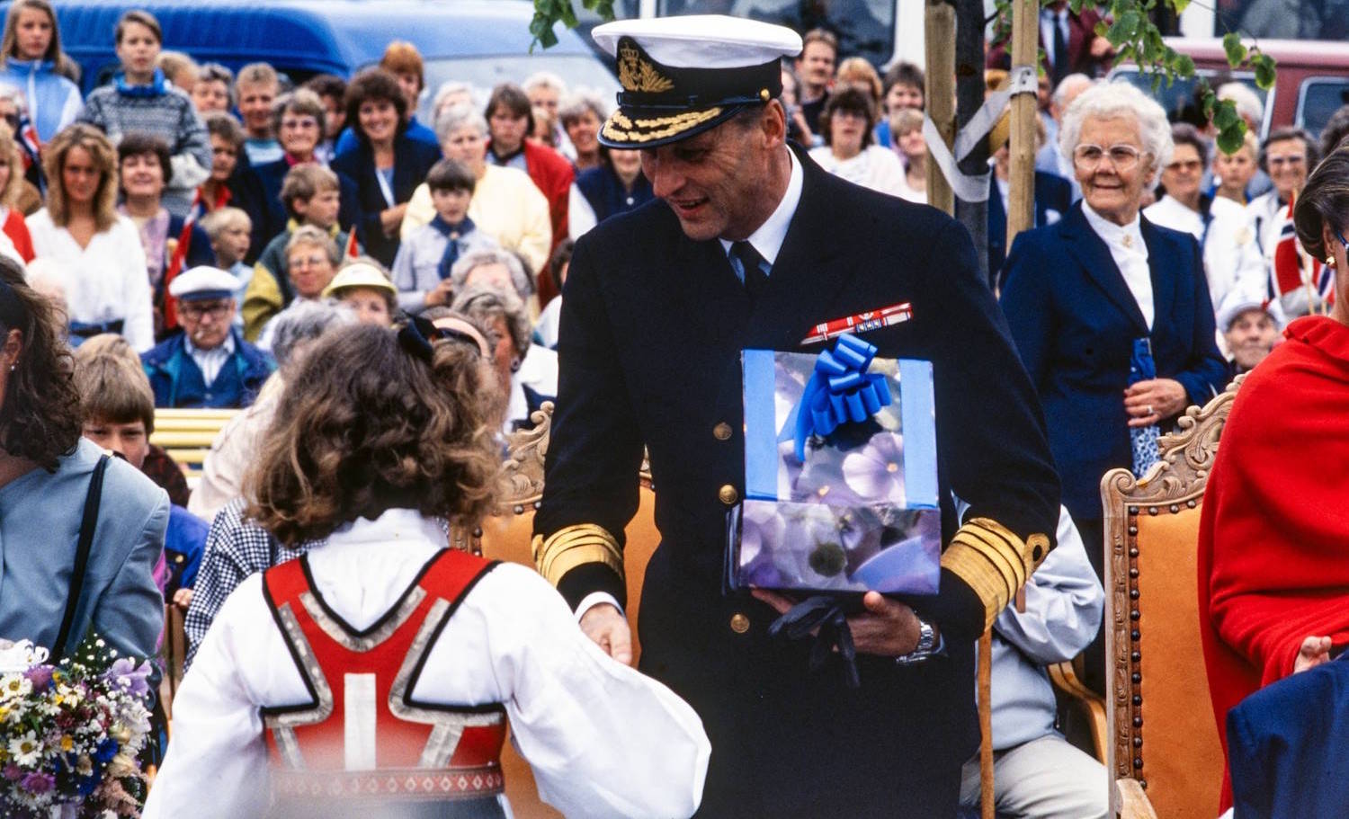 Kong Harald 5. mottar gave under signingsferden i Grimstad 28.06.1991. Foto Erik Holand, Agderposten. Bildet viser en offentlig markering utendørs med et stort publikum samlet i bakgrunnen. I forgrunnen står en person i mørk marineuniform med hvit uniformslue og gullfargede detaljer på ermene. Personen bøyer seg frem og tar imot en innpakket gave med blå sløyfe fra et barn som står med ryggen mot kameraet og er kledd i bunad. Rundt dem står flere personer på og ved stoler med utskårne tredetaljer. Til høyre i bildet sees en person innhyllet i et sterkt rødt sjal eller poncho, mens en eldre person i mørk blå drakt står litt lenger bak. En blomsterbukett med lilla, hvite og gule blomster ligger nederst til venstre i bildet. Bakgrunnen er fylt av mennesker i ulike aldre som følger seremonien. Flere små norske flagg er synlige blant publikum. Et tre med grønne blader strekker seg inn over øvre del av bildet, og parkerte kjøretøy kan skimtes bak folkemengden. Bildet formidler en høytidelig gaveoverrekkelse under et offisielt arrangement.