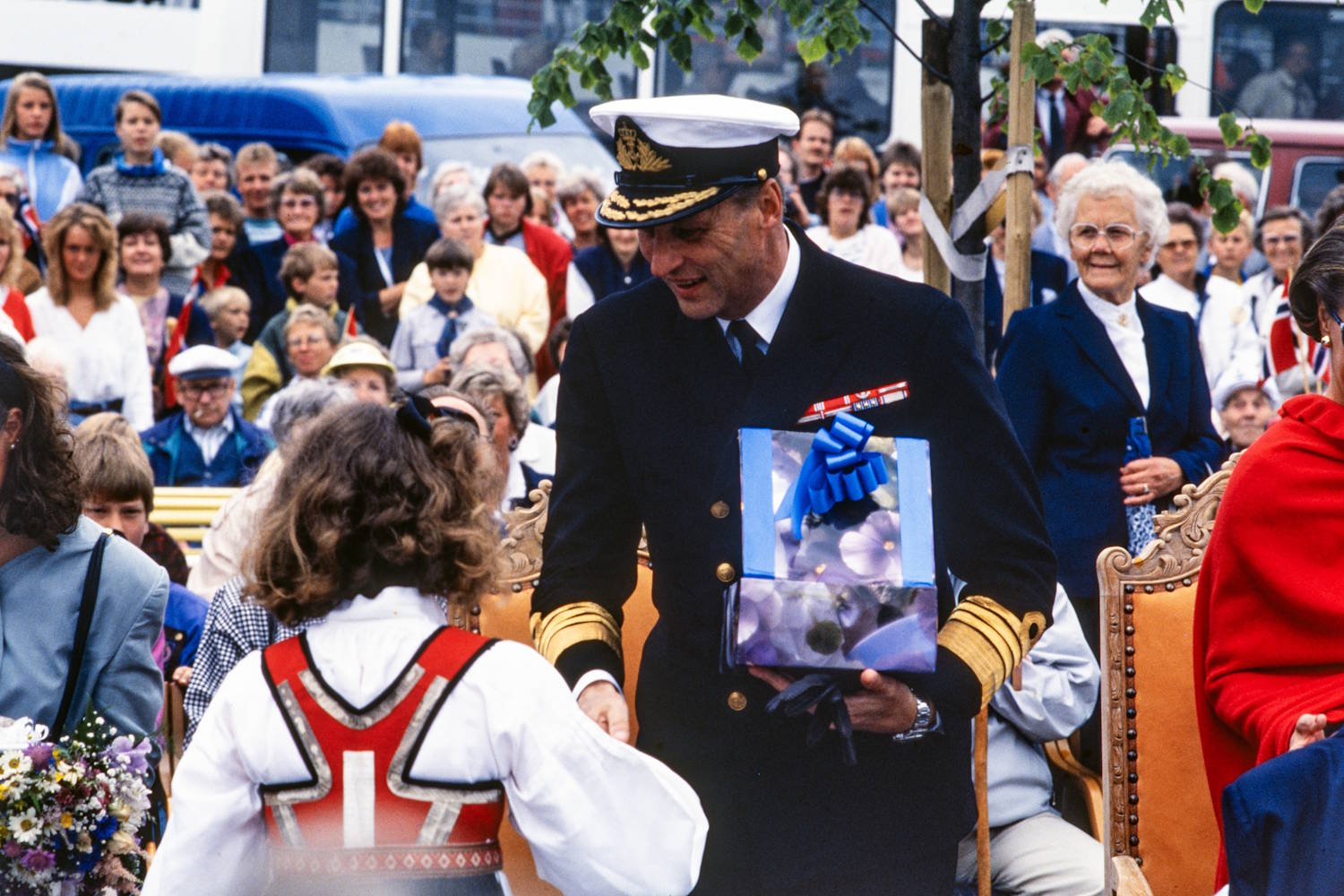 Kong Harald 5. mottar gave under signingsferden i Grimstad 28.06.1991. Foto Erik Holand, Agderposten. Bildet viser en offentlig markering utendørs med et stort publikum samlet i bakgrunnen. I forgrunnen står en person i mørk marineuniform med hvit uniformslue og gullfargede detaljer på ermene. Personen bøyer seg frem og tar imot en innpakket gave med blå sløyfe fra et barn som står med ryggen mot kameraet og er kledd i bunad. Rundt dem står flere personer på og ved stoler med utskårne tredetaljer. Til høyre i bildet sees en person innhyllet i et sterkt rødt sjal eller poncho, mens en eldre person i mørk blå drakt står litt lenger bak. En blomsterbukett med lilla, hvite og gule blomster ligger nederst til venstre i bildet. Bakgrunnen er fylt av mennesker i ulike aldre som følger seremonien. Flere små norske flagg er synlige blant publikum. Et tre med grønne blader strekker seg inn over øvre del av bildet, og parkerte kjøretøy kan skimtes bak folkemengden. Bildet formidler en høytidelig gaveoverrekkelse under et offisielt arrangement.