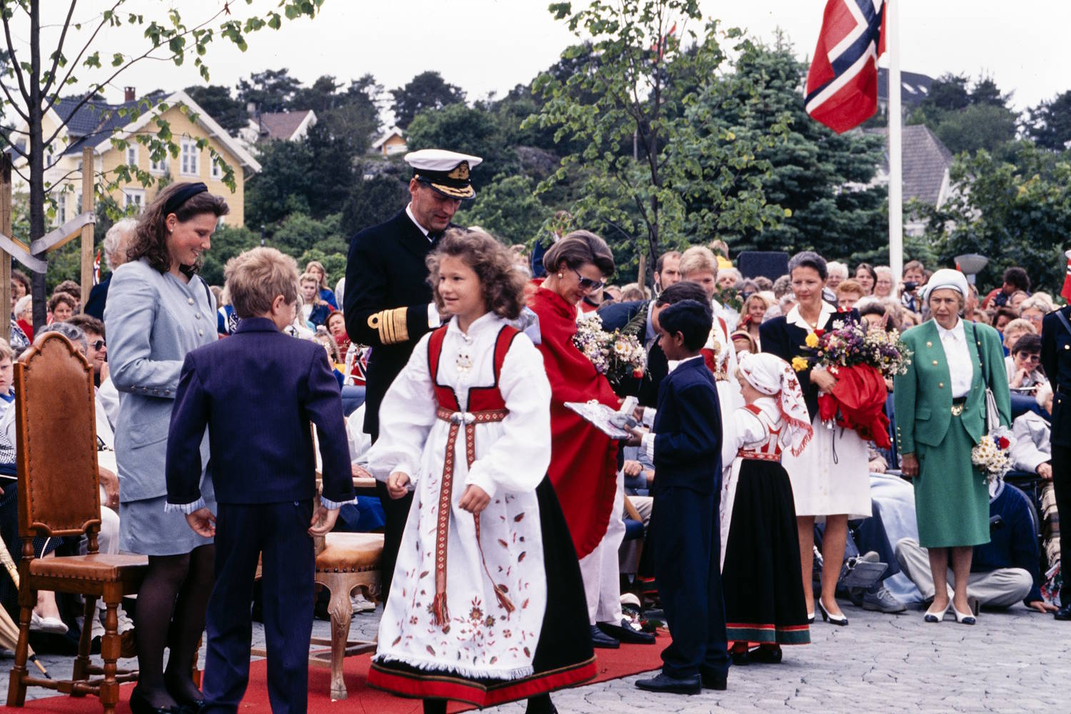 Kongefamilien mottar gaver under signingsferden i Grimstad 28.06.1991. Foto Erik Holand, Agderposten. Bildet viser en utendørs seremoni på en plass med brosteinsdekke, omgitt av et stort publikum. I forgrunnen står flere barn i bunader og høytidsklær foran en gruppe voksne som mottar blomster og gaver. Midt i bildet står en person i mørk marineuniform med hvit uniformslue. Ved siden av står en person innhyllet i et sterkt rødt sjal og holder en blomsterbukett. Flere barn er samlet rundt dem, blant annet en jente i hvit og rød bunad som går mot kameraet. Et annet barn holder fram en gave eller et brett. Til venstre står en person i lys blå drakt ved siden av en gutt i mørk dress. Flere utskårne trestoler er plassert på et rødt teppe. Til høyre står en person i grønn drakt, mens andre deltakere holder store blomsterbuketter. Bakgrunnen er fylt av tilskuere, norske flagg og grønne trær. Mellom trærne kan en se lyse bolighus. Fargene i bildet preges av rødt, hvitt, blått og grønt. Bildet formidler en høytidelig gave- og blomsteroverrekkelse under et offisielt besøk.