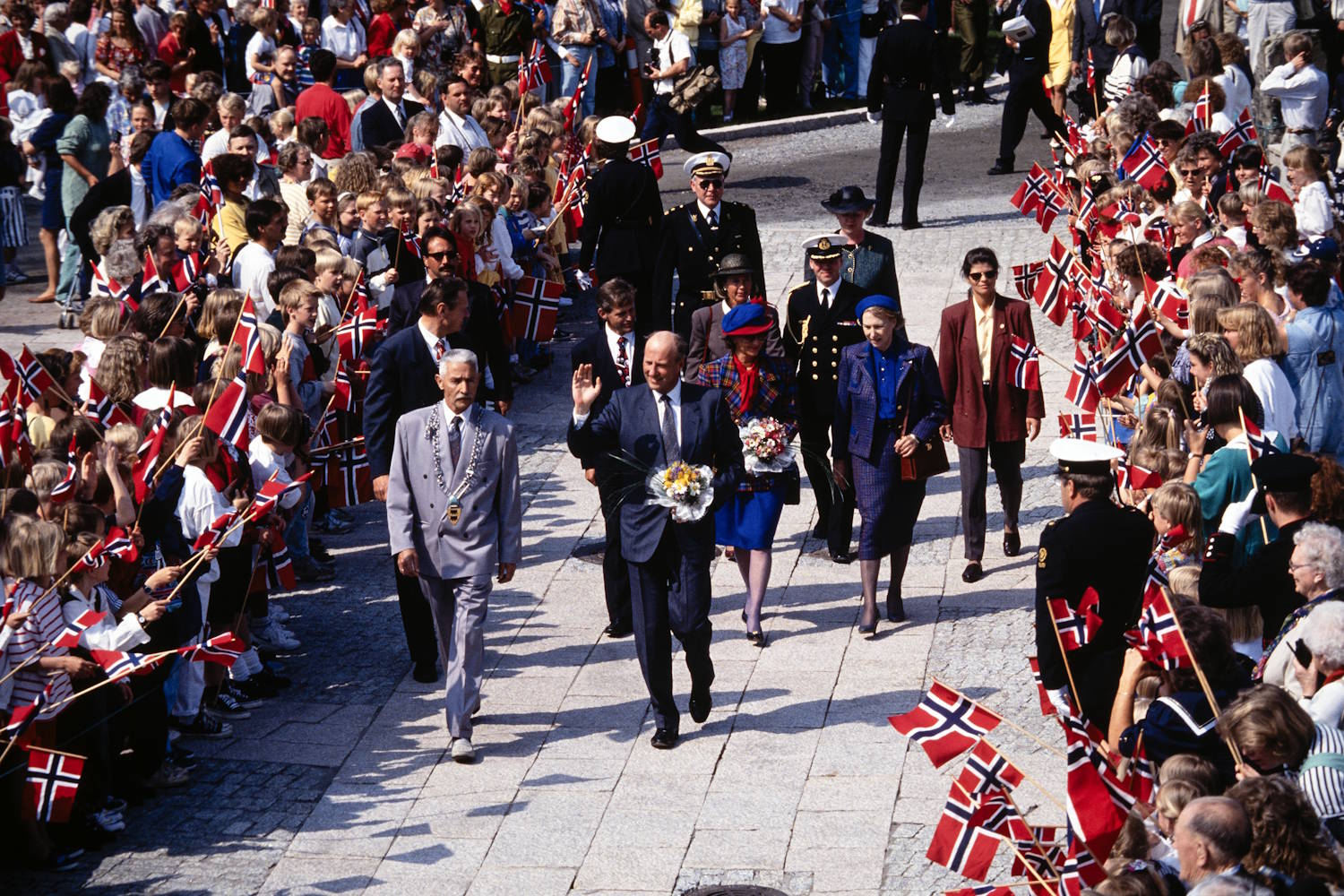 Kongeparet ankommer Setesdal Mineralpark 10. mai 1993. Foto: Arild Jakobsen, Fædrelandsvennen. Bildet viser en stor folkemengde samlet langs en bred, steinlagt gate eller plass. På begge sider står mennesker tett i tett og vifter med små norske flagg. I midten av bildet går et følge med flere personer omgitt av uniformert personell. Foran i følget går en person i lys grå dress med et kjede rundt halsen. Like bak følger flere personer i mørke dresser og fargerike antrekk. En av personene i sentrum holder en blomsterbukett og løfter hånden. Flere uniformerte vakter er synlige både i følget og langs ruten. Bildet er tatt ovenfra, noe som gir oversikt over den lange rekken av mennesker og flagg som danner en ramme rundt følget. Fargene domineres av rødt, hvitt og blått fra de mange norske flaggene. Solskinn og skarpe skygger tyder på klart vær. Stemningen fremstår som høytidelig og preget av en offentlig markering eller seremoni med stor publikumsoppmøte.