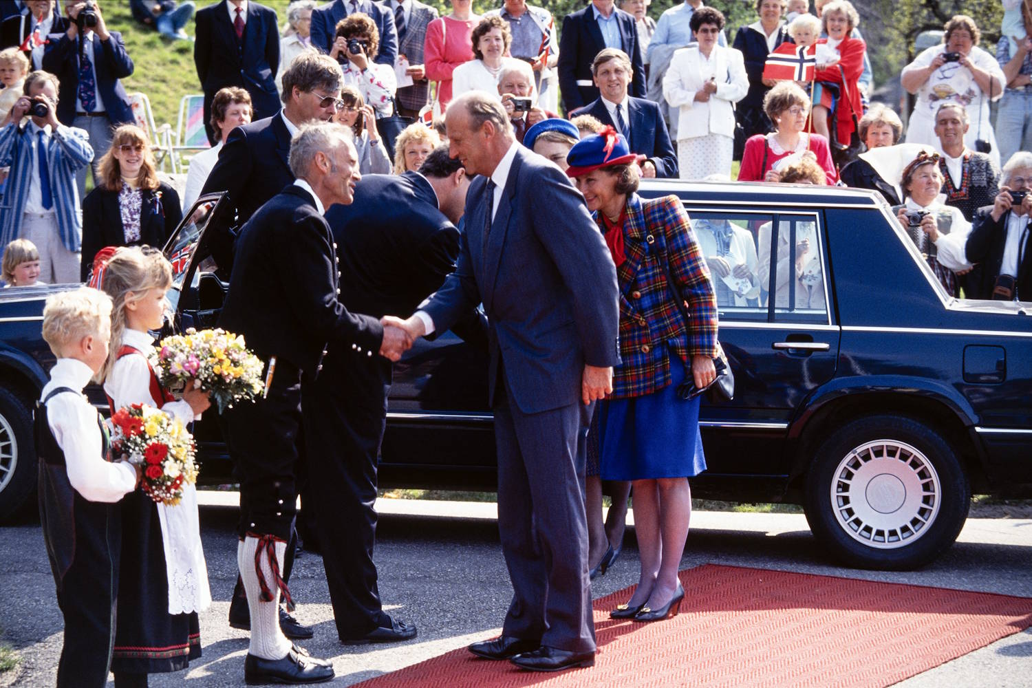 Kongeparet ønskes velkommen ved kommunehuset i Iveland 10. mai 1993. Foto: Kjartan Bjelland, Fædrelandsvennen. Bildet viser en høytidelig velkomstseremoni utendørs. En mørk personbil står parkert ved en rød løper, omgitt av mange mennesker. I forgrunnen håndhilser to personer på hverandre ved siden av bilen. Ved siden av står en person i rutete jakke, blått skjørt og blå hatt med røde detaljer. Foran velkomstfølget står to barn i tradisjonelle klær og holder hver sin blomsterbukett. Rundt dem står flere voksne, noen i bunad og andre i dress. I bakgrunnen er det samlet mange tilskuere. Flere holder norske flagg, og noen fotograferer eller filmer hendelsen. Sollyset er sterkt og gir klare skygger på bakken. Bildet formidler en formell mottakelse med stor lokal oppslutning, der gjester ønskes velkommen med blomster, flagg og tradisjonelle innslag.