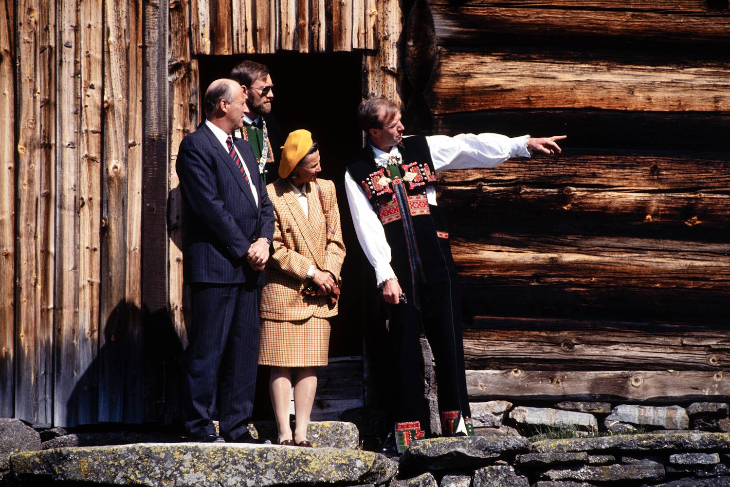 Kongeparet på Rygnestadtunet 11. mai 1993. Foto: Arild Jakobsen, Fædrelandsvennen. Bildet viser fire personer som står foran en eldre tømmerbygning. Bygningen er oppført i grovt, mørkt tømmer med værslitte vegger, og en åpen dør danner en mørk bakgrunn midt i motivet. To personer står fremst på en steinmur eller steinlagt avsats. Den ene er kledd i mørk dress med slips. Ved siden av står en person i en lysrutet drakt og en gul hatt. Bak dem står en tredje person med solbriller og grønn vest under en mørk jakke. Til høyre står en person i tradisjonsdrakt med rikt broderte detaljer på vesten. Personen strekker ut armen og peker mot høyre, som om noe forklares eller vises fram til de andre. Sterkt sollys faller på bygningen og personene, og skaper tydelige skygger. Fargene domineres av varme bruntoner fra tømmerveggen, kombinert med blått, gult, grønt og rødt i klærne. Foran bygningen ligger store steiner med flekker av lav. Motivet framstår som et besøk ved et historisk tun eller kulturminne, der en omvisning eller presentasjon pågår utendørs.