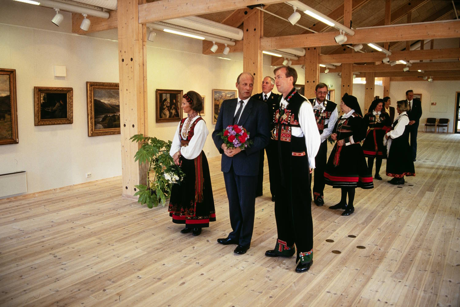 Kongeparet under åpningen av Setesdalsmuseets nybygg på Rysstad 10. mai 1993. Foto: Kjartan Bjelland, Fædrelandsvennen. Bildet viser et lyst og romslig museums- eller gallerirom med tregulv, synlige trebjelker i taket og malerier hengende på veggene. Flere personer er samlet i en gruppe midt i salen. Foran står en person i mørk dress som holder en bukett med røde og lilla blomster. Ved siden av står en person i tradisjonell folkedrakt med rikt broderte detaljer i rødt, grønt og hvitt. Flere andre personer i lignende drakter står litt bakover i rommet. En uniformert person er også synlig i bakgrunnen. Gruppen ser ut til å bevege seg gjennom utstillingen mens de betrakter kunsten på veggene. Til venstre står en grønn potteplante ved en av de kraftige trestolpene som bærer taket. Rommet er preget av lyse treflater, varme fargetoner og jevn belysning fra takmonterte lamper. Motivet framstår som en formell omvisning eller åpning av en kultur- eller museumsutstilling.